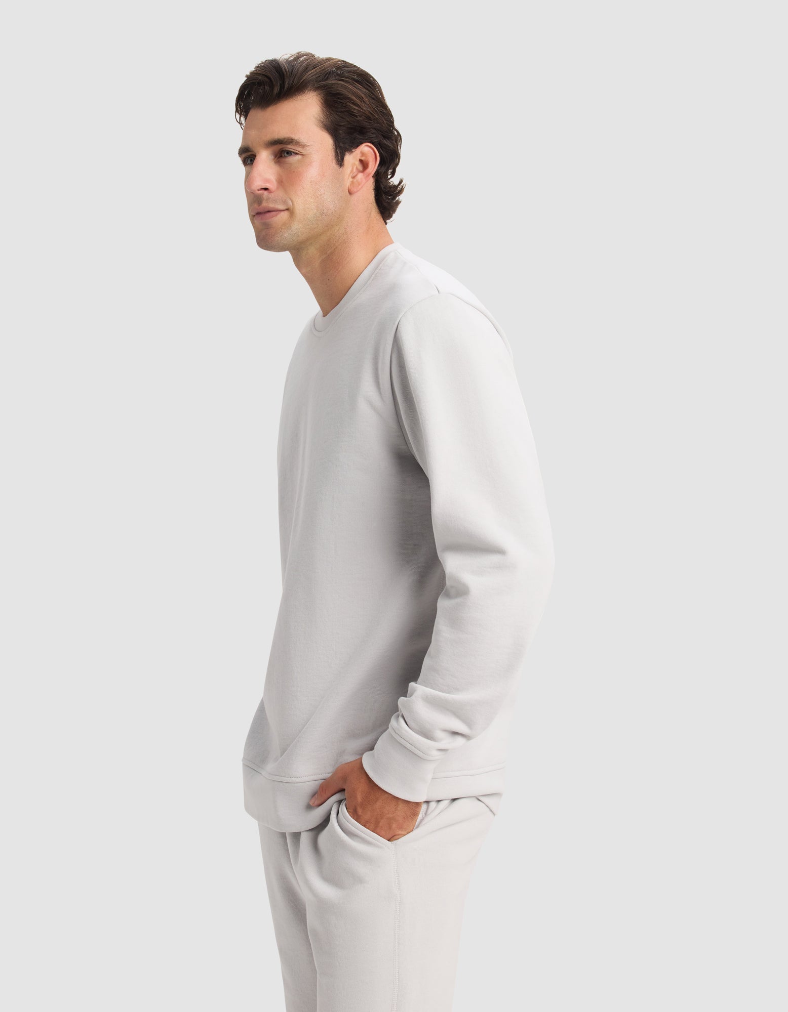 A man with short brown hair wears the Men's Dylan Crewneck from Cozy Earth with matching pants, standing sideways with hands in pockets, looking ahead against a plain light gray background.