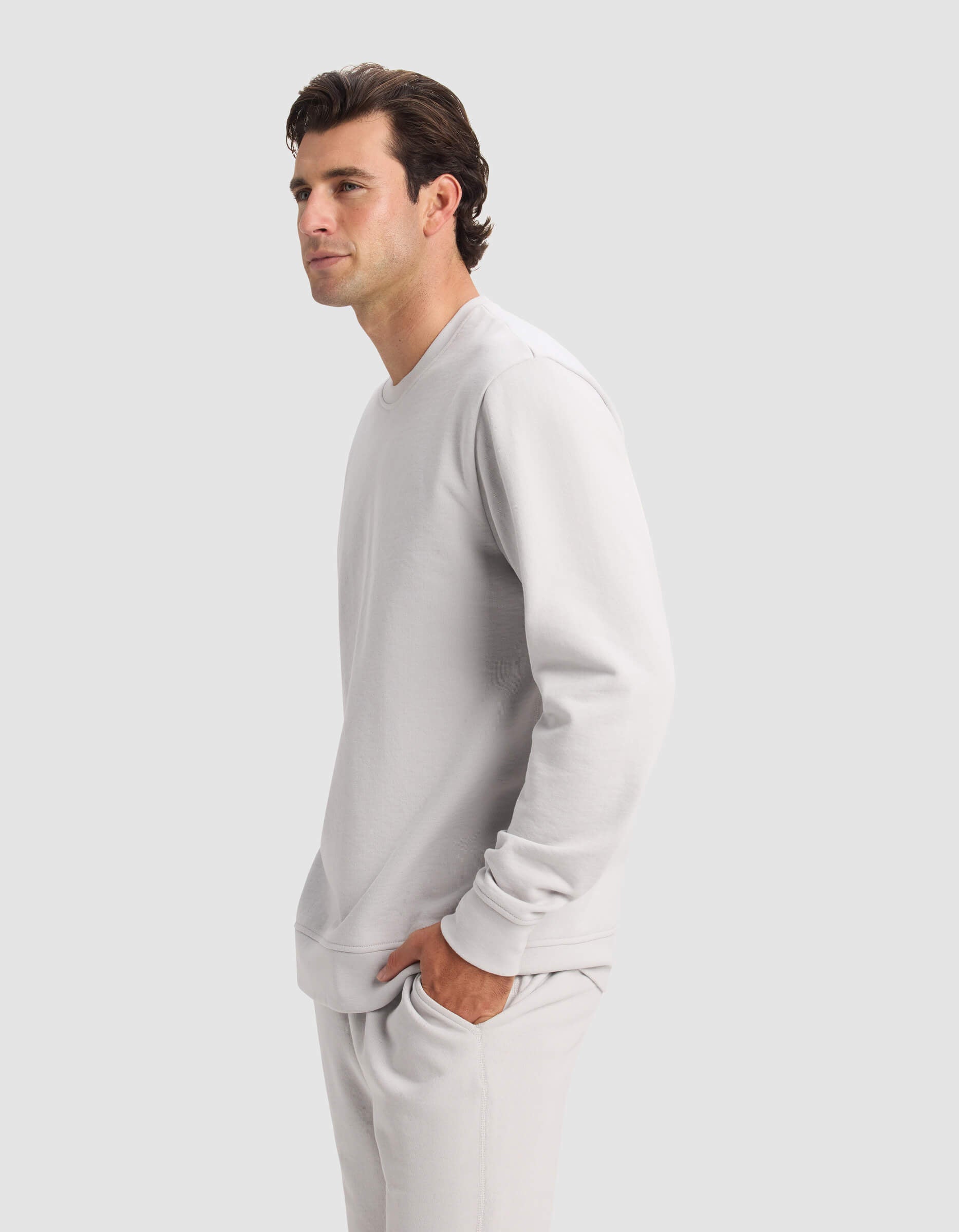 A man with dark hair models the Cozy Earth Men's Dylan Crewneck and matching pants in light gray, standing in profile with hands in his pockets against a plain light gray background.