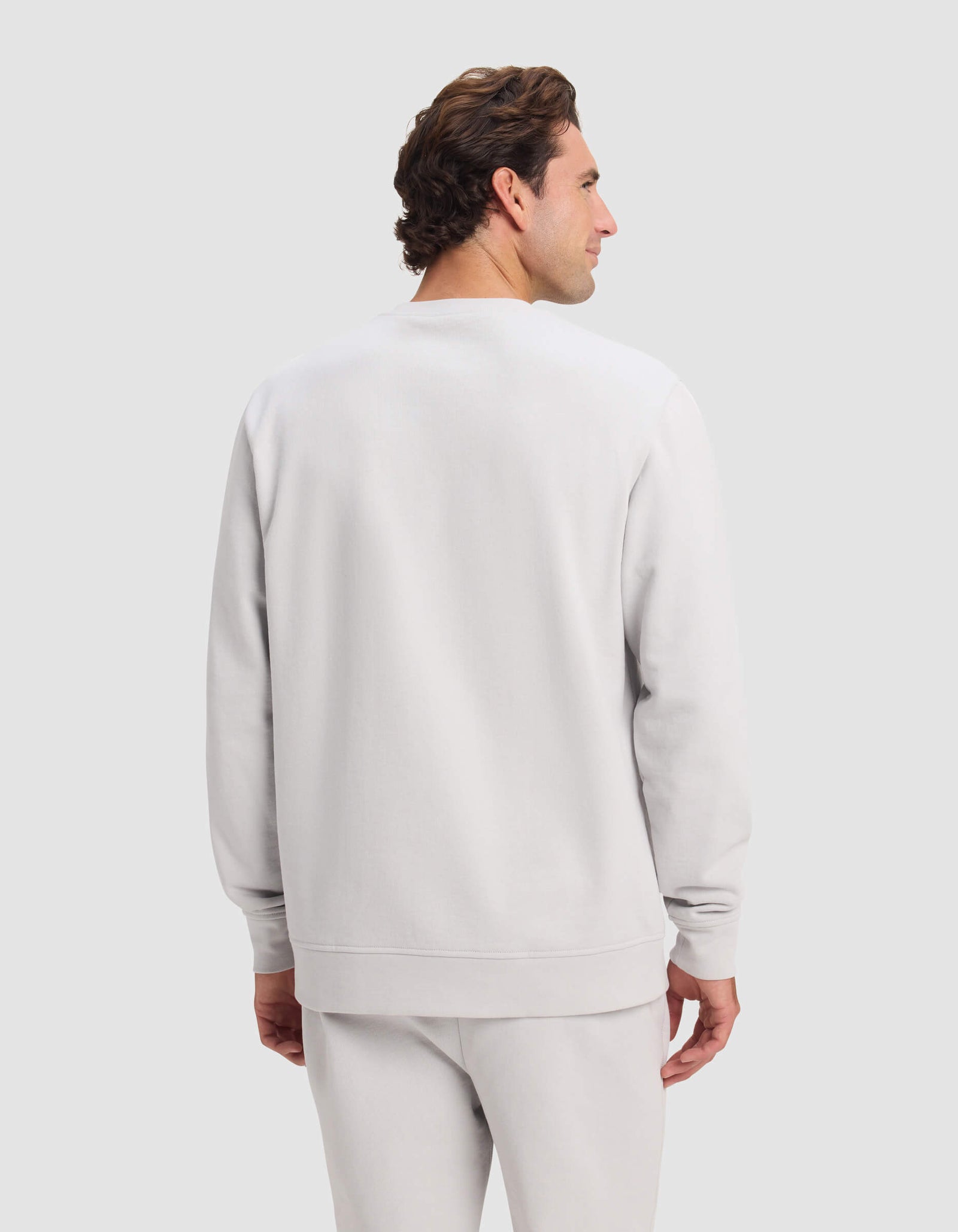 A man with short brown hair is seen from the back, wearing Cozy Earth's Men's Dylan Crewneck sweatshirt and matching sweatpants, standing against a light gray background.