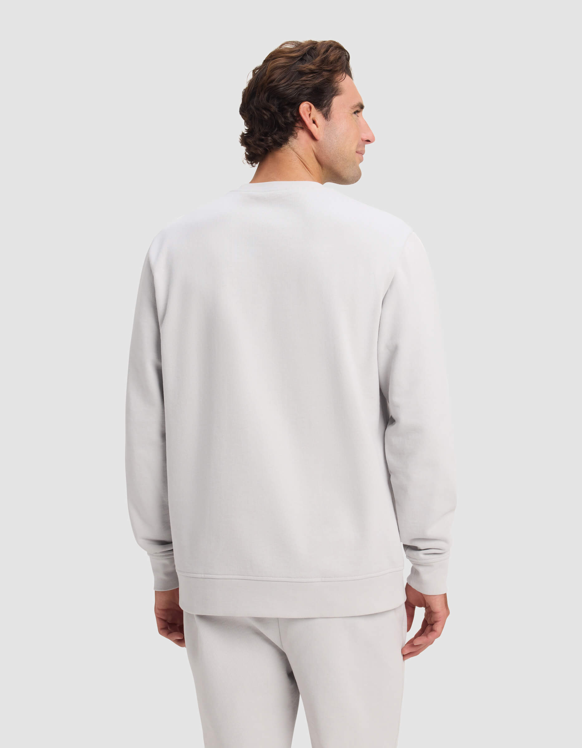 A man with short brown hair is seen from the back, wearing Cozy Earth's Men's Dylan Crewneck sweatshirt and matching sweatpants, standing against a light gray background.