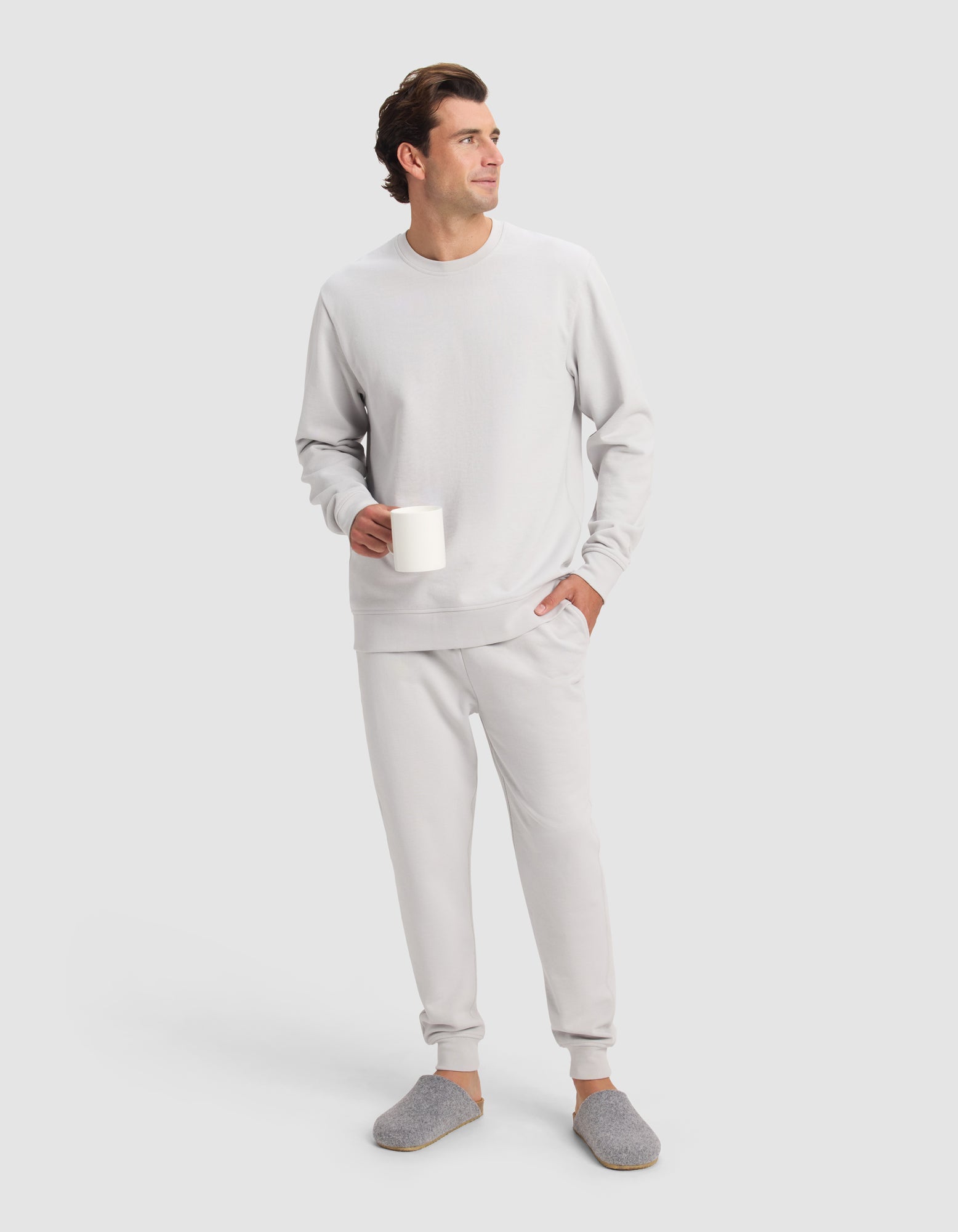 A man in the Cozy Earth Men's Dylan Crewneck and matching joggers stands holding a white mug, wearing gray slippers and looking off to the side against a plain light background.
