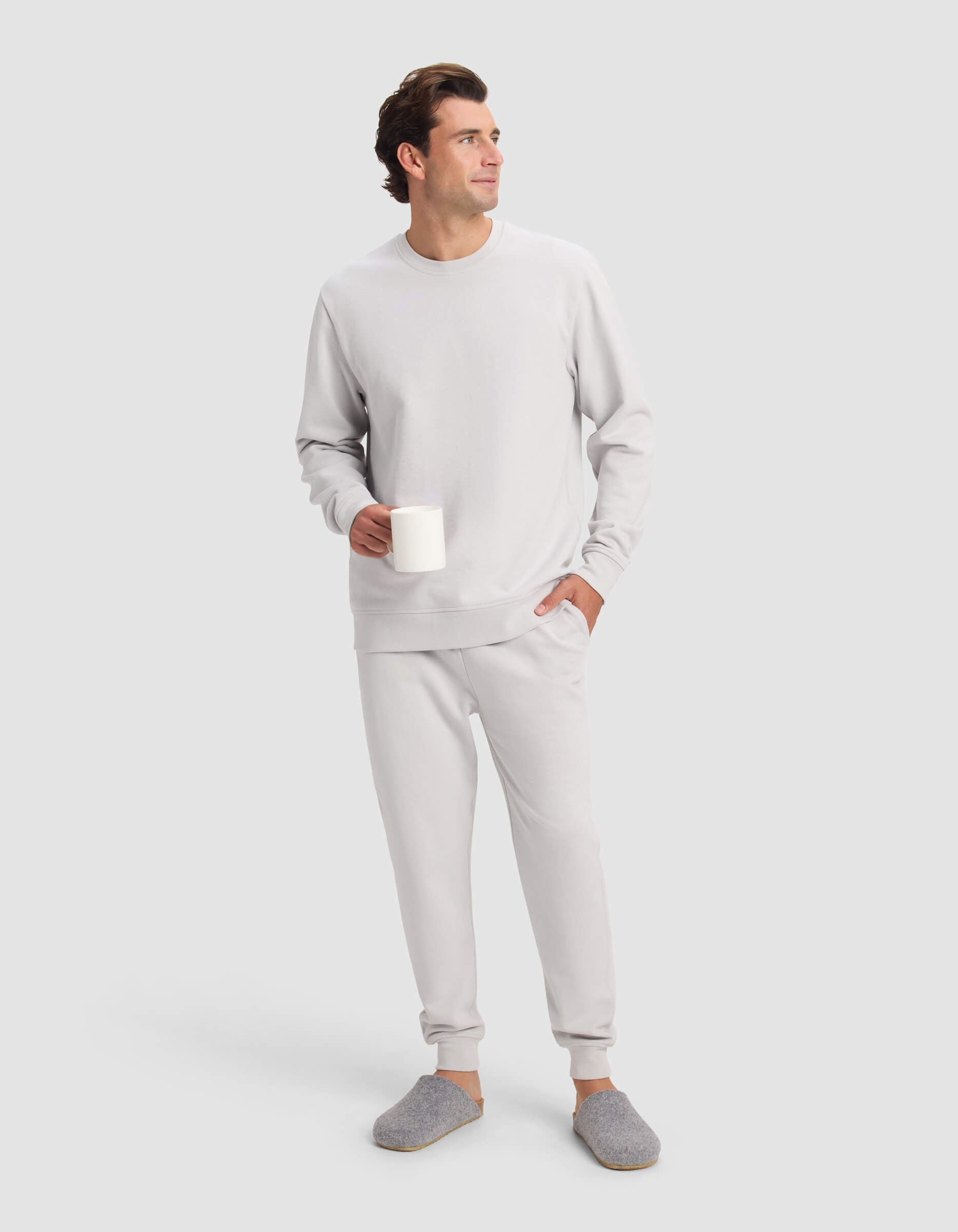 A man wearing the Cozy Earth Men's Dylan Crewneck and gray slippers stands holding a white mug, looking off to the side against a plain light background.