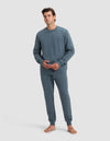 A man with short dark hair, barefoot against a plain background, wears the Cozy Earth Men's Dylan Crewneck & Jogger Set in blue-gray, hands lightly clasped, and maintains a neutral expression.|Color:Dawn
