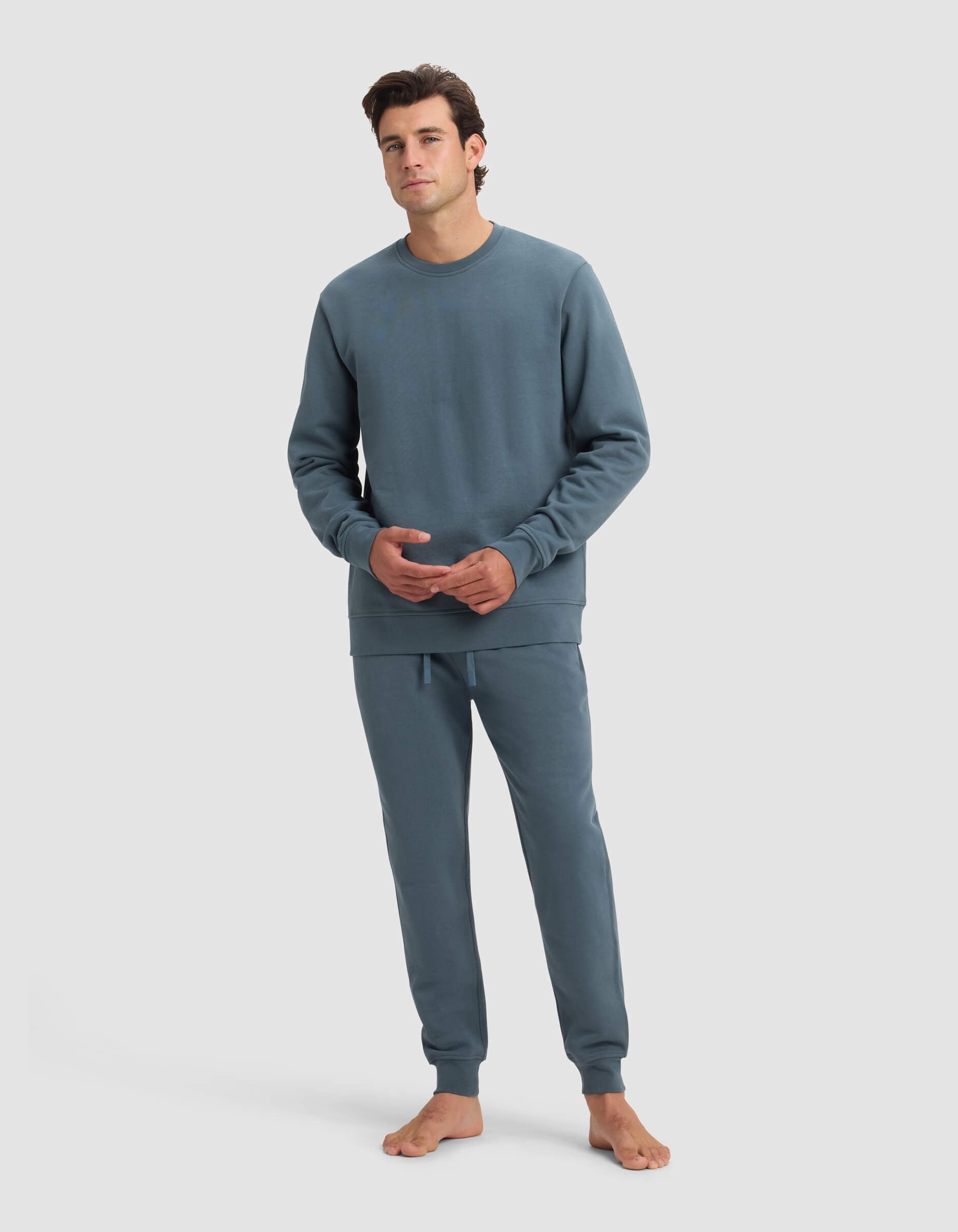 A man with short dark hair, barefoot against a plain background, wears the Cozy Earth Men's Dylan Crewneck & Jogger Set in blue-gray, hands lightly clasped, and maintains a neutral expression.