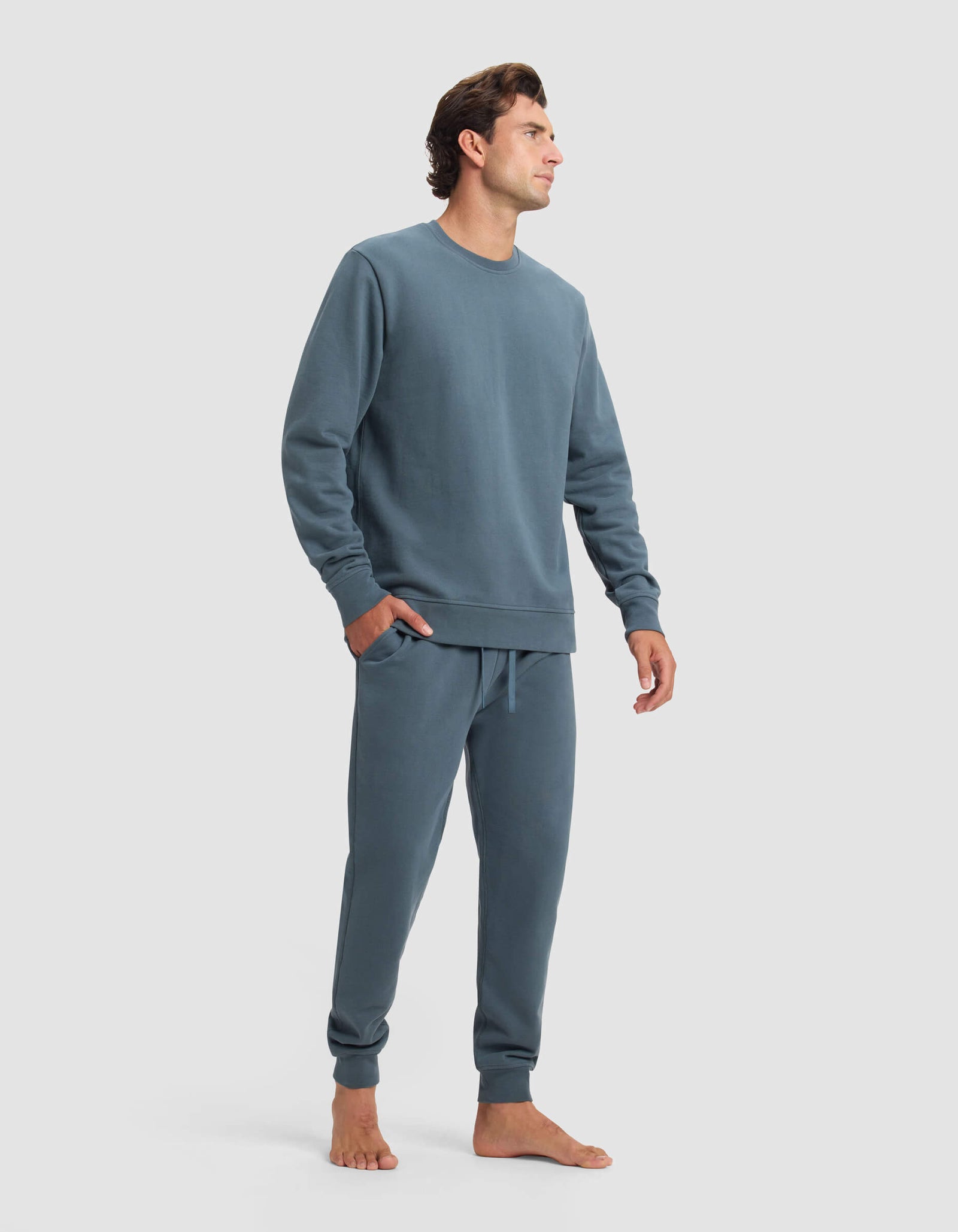 A man stands barefoot against a plain light gray background, wearing the Cozy Earth Men's Dylan Crewneck & Jogger Set in blue-gray, with his left hand in his pocket and looking off to the side.