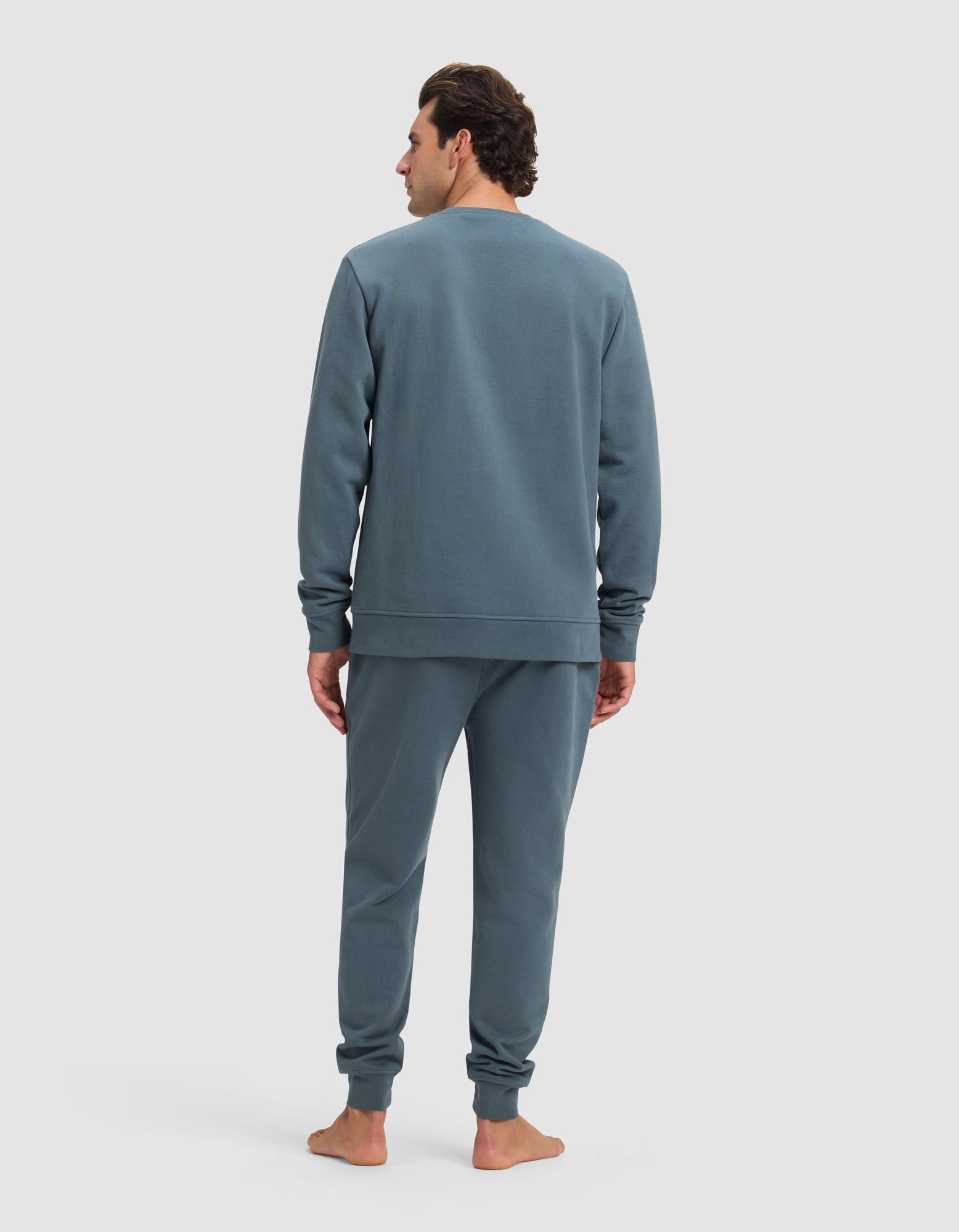 A man stands barefoot facing away, wearing the Cozy Earth Men's Dylan Crewneck & Jogger Set in blue-gray, set against a plain light background.