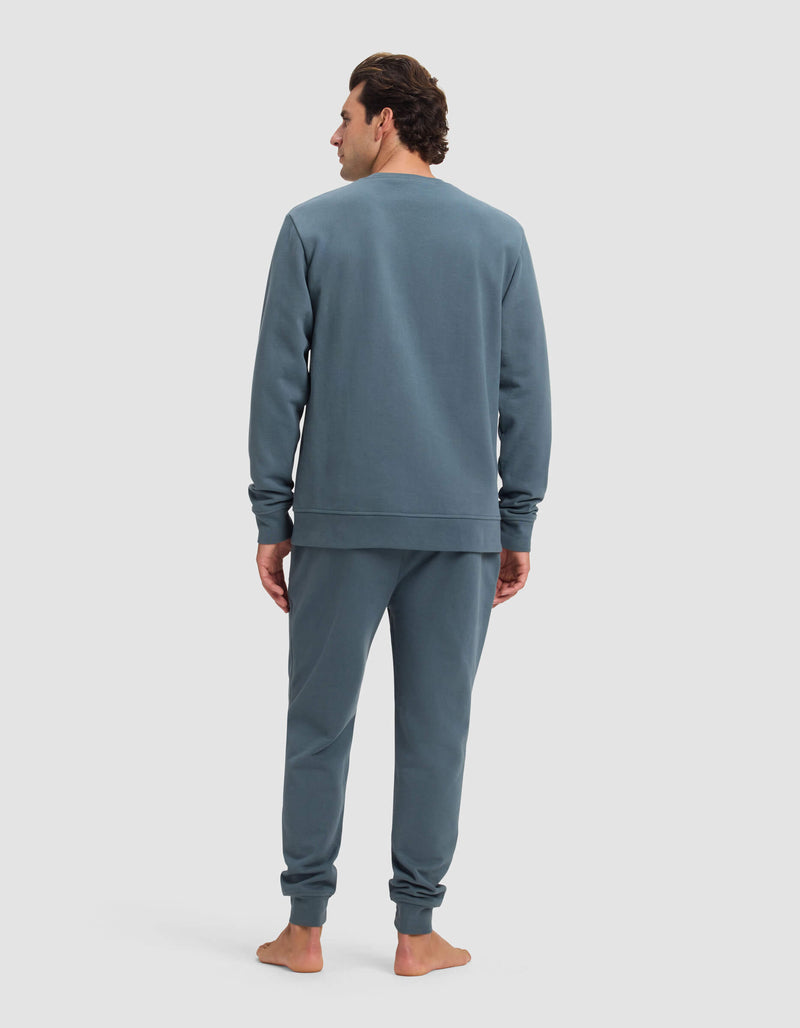A man stands barefoot facing away, wearing the Cozy Earth Men's Dylan Crewneck & Jogger Set in blue-gray against a plain light background.
