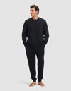 A man stands barefoot against a plain background, wearing the Cozy Earth Men's Dylan Crewneck & Jogger Set in black. With his hands in his pockets, he looks slightly to the side and has a relaxed expression.|Color:Jet Black