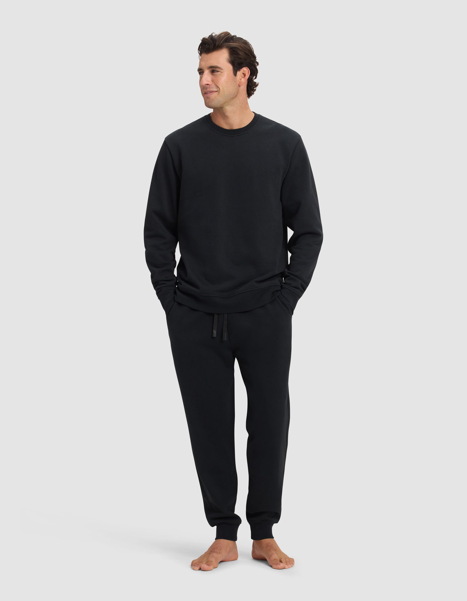 A man stands barefoot against a plain background, wearing the Cozy Earth Men's Dylan Crewneck & Jogger Set in black. With his hands in his pockets, he looks slightly to the side and has a relaxed expression.|Color:Jet Black