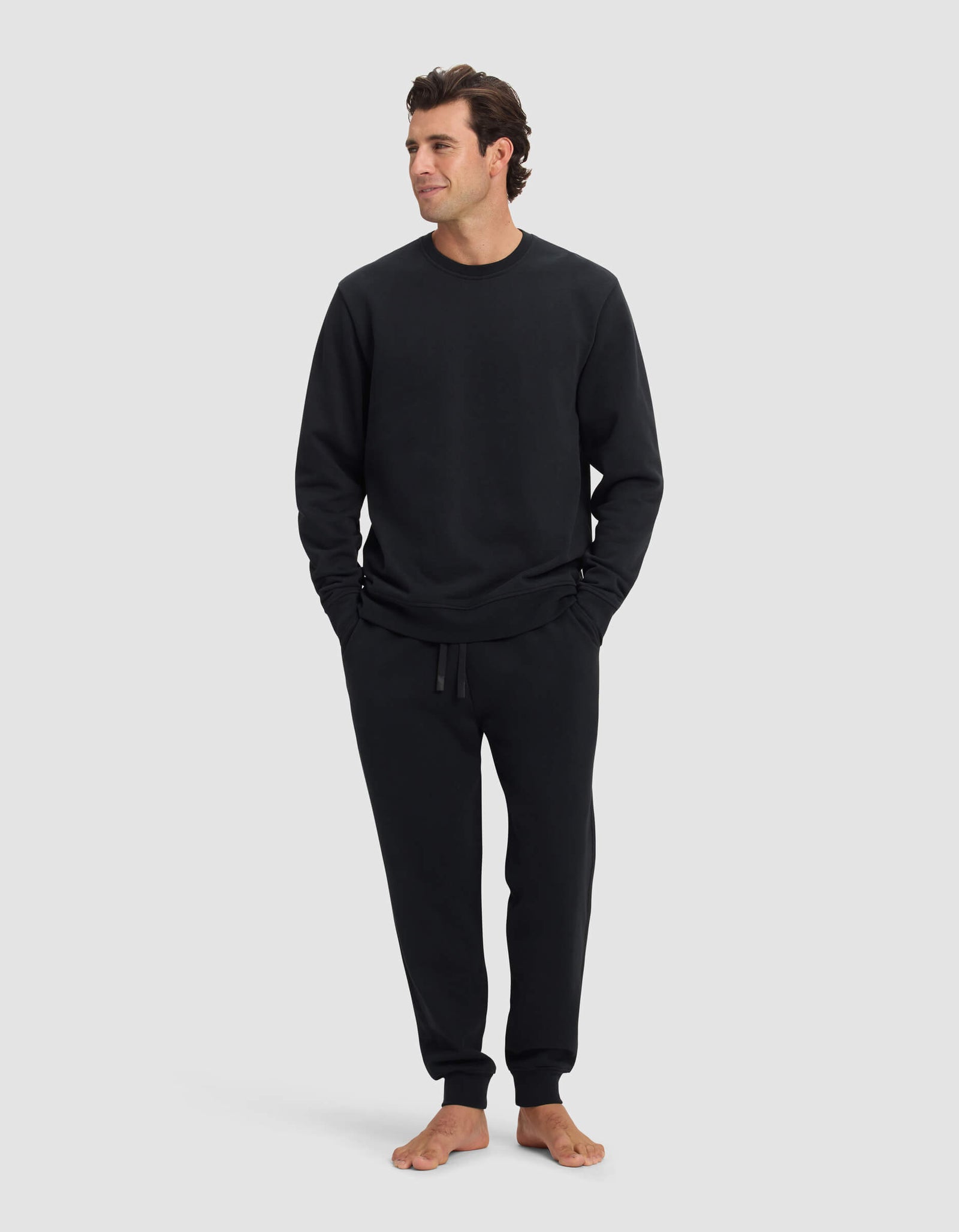 A man stands barefoot against a light gray background, wearing the Cozy Earth Men's Dylan Crewneck & Jogger Set in black. He has his hands in his pockets and gazes off to the side with a relaxed expression.