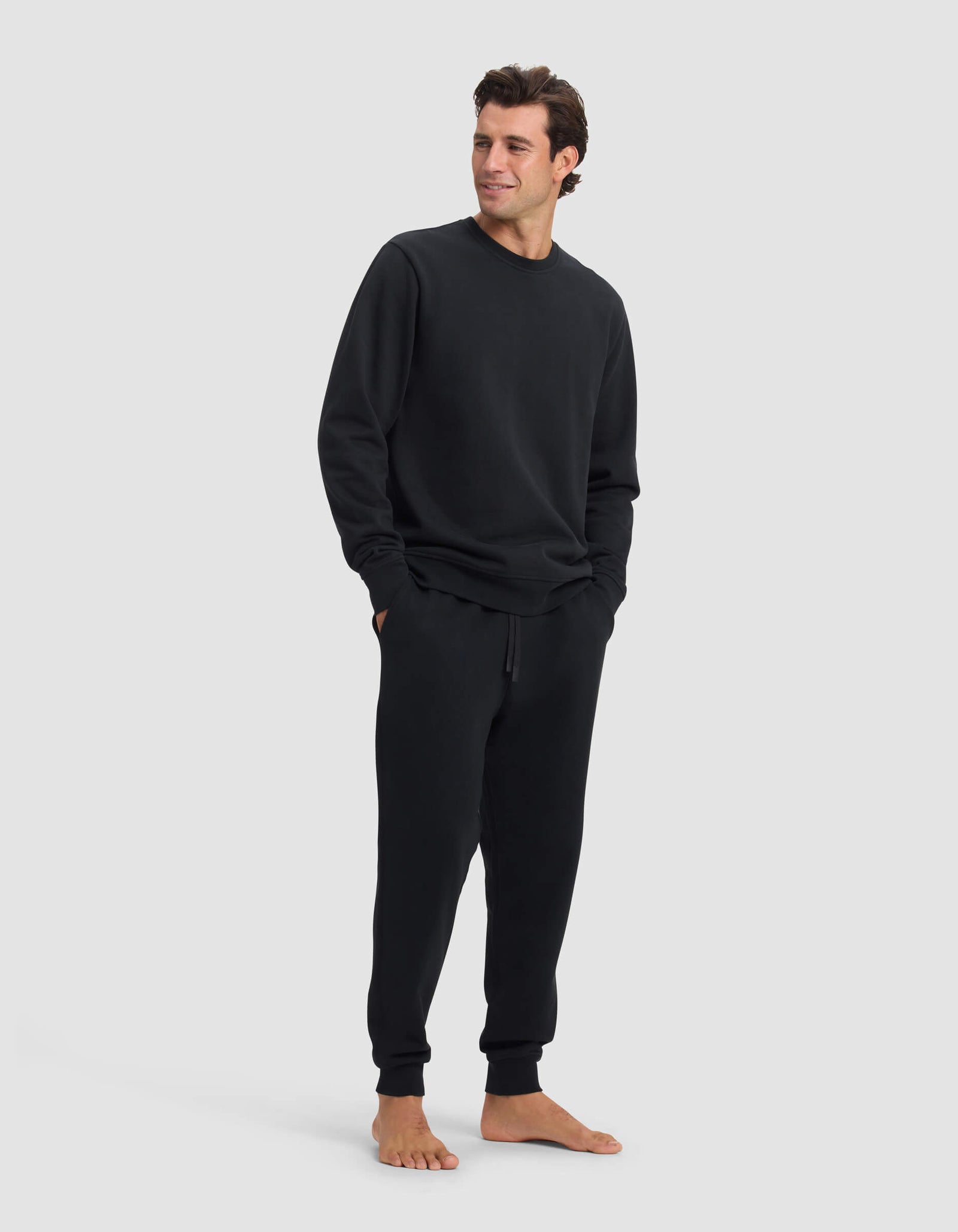 A man stands barefoot against a plain background, wearing the Cozy Earth Men's Dylan Crewneck & Jogger Set in black. He has his hands in his pockets and looks slightly to the side with a slight smile.