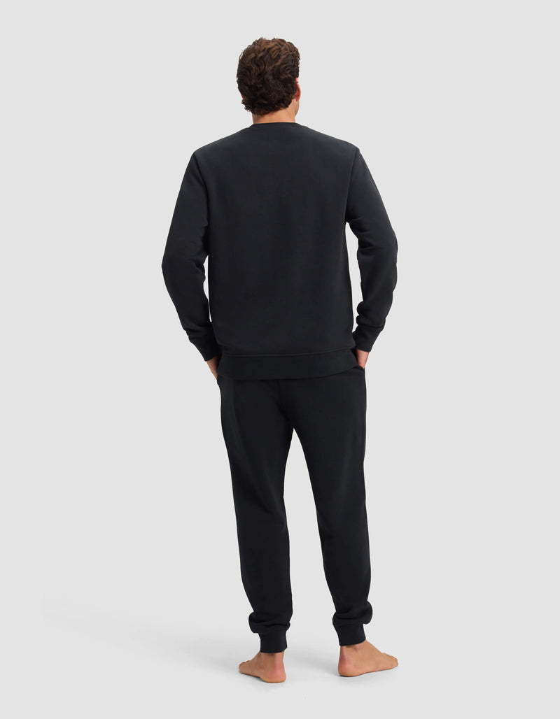 A person stands barefoot, facing away, wearing the Men's Dylan Crewneck & Jogger Set by Cozy Earth in black. Their hands are in their pockets against a plain light gray background.