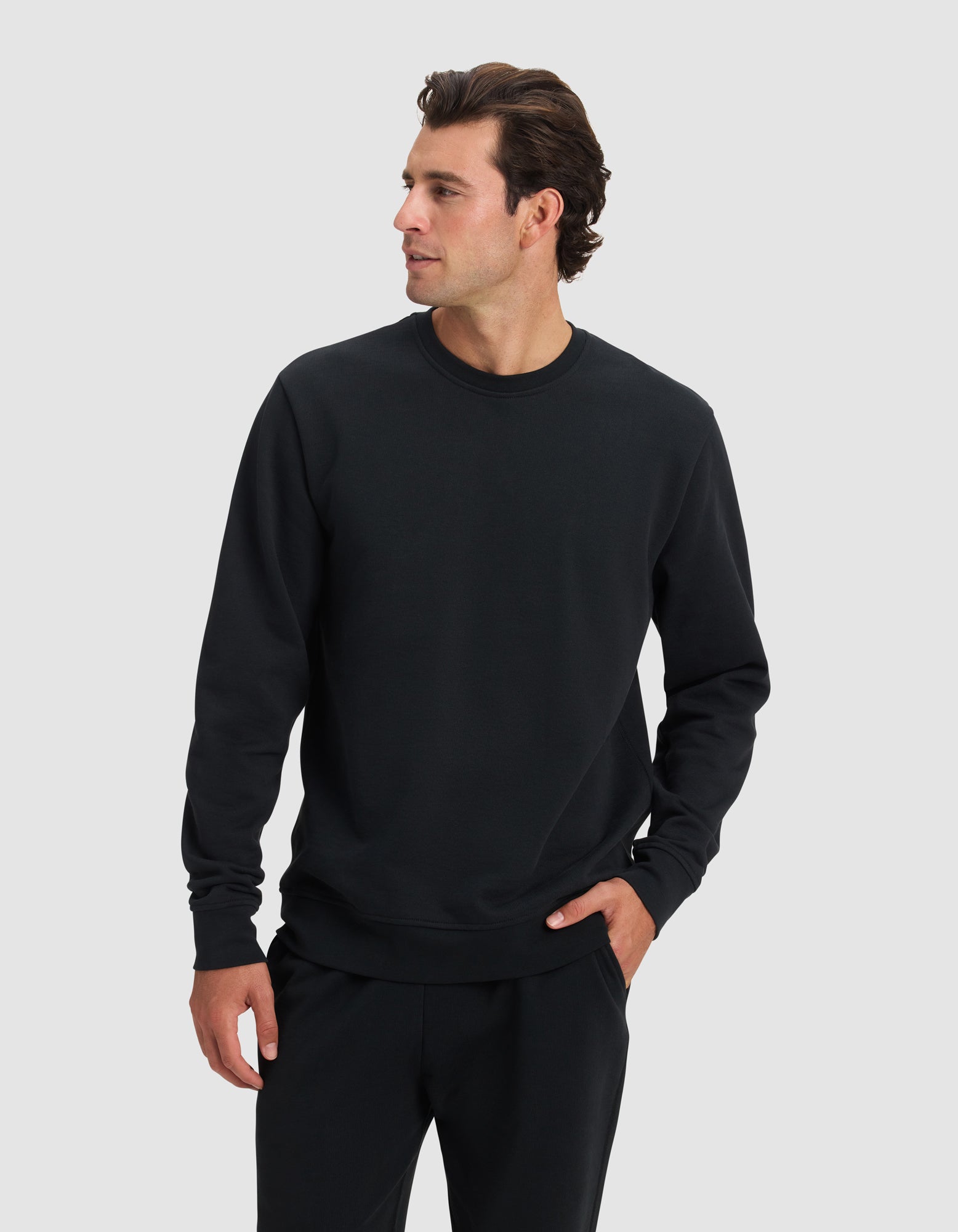 A man models the Cozy Earth Men's Dylan Crewneck and matching black pants, standing against a light gray background with one hand in his pocket and gazing to his right.