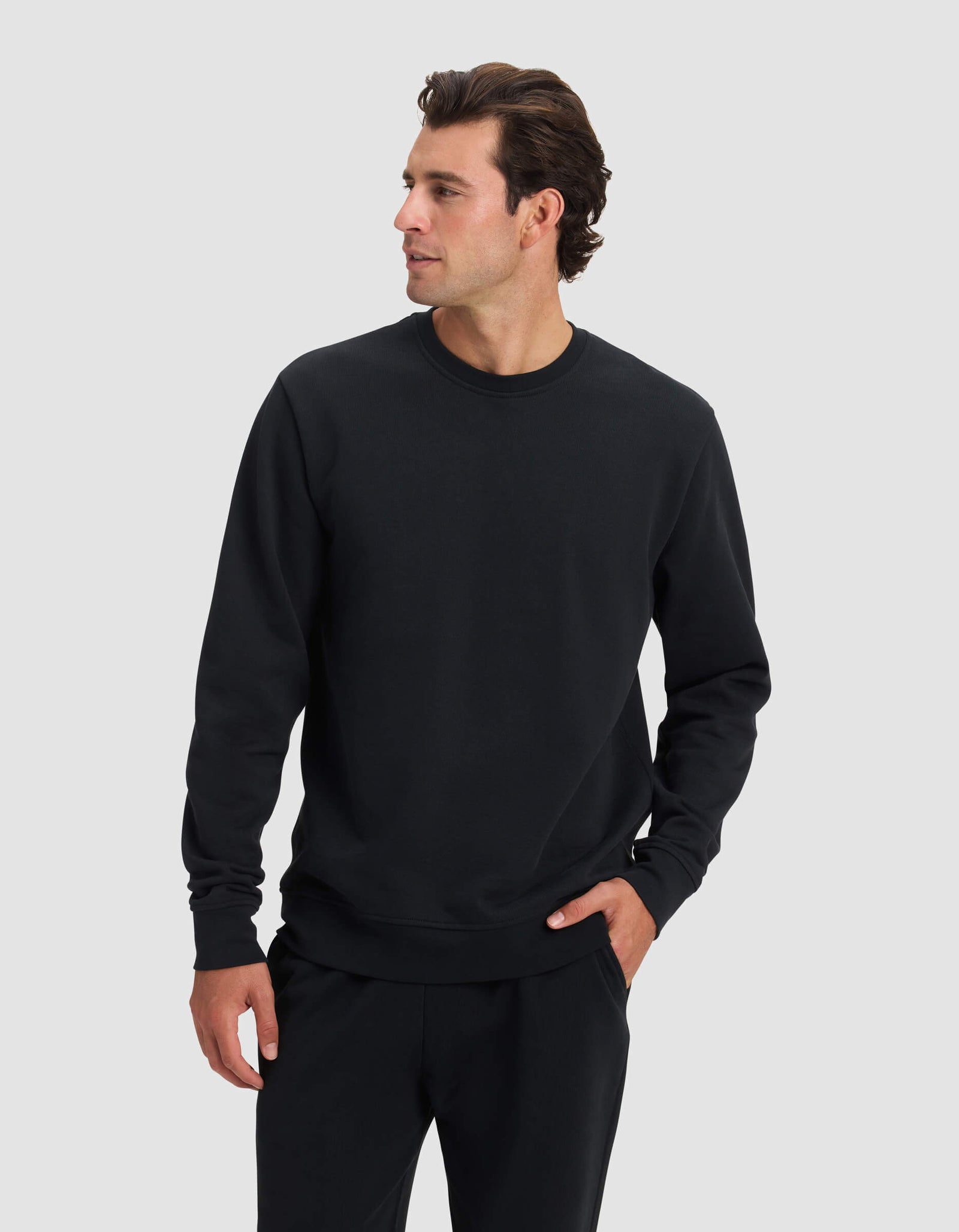 A man wearing the Cozy Earth Men's Dylan Crewneck in black and matching black pants stands against a light gray background, looking to his left with one hand in his pocket.