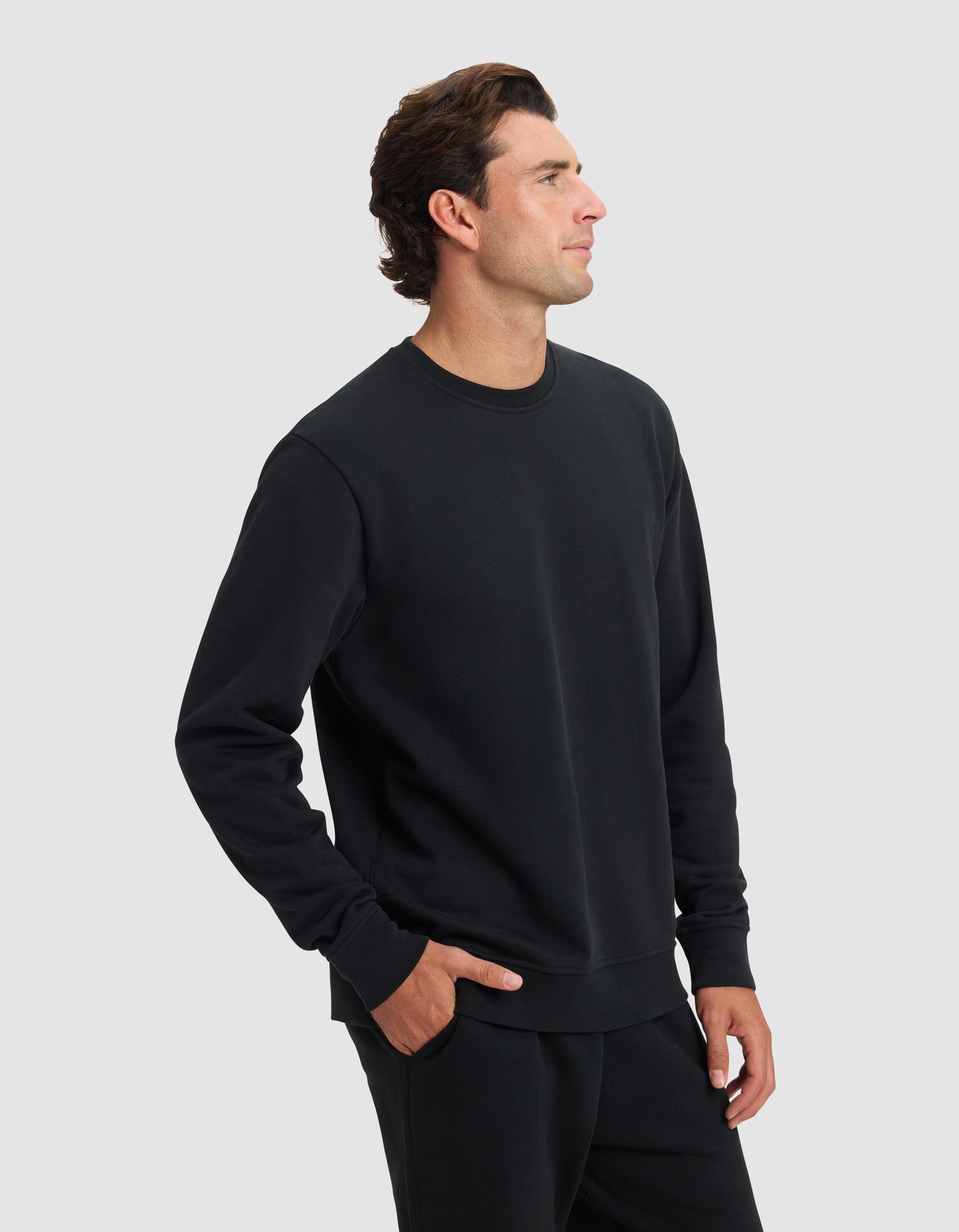 A man stands against a light gray background wearing the Cozy Earth Men's Dylan Crewneck and matching black pants, one hand in his pocket and looking to the side with a neutral expression.