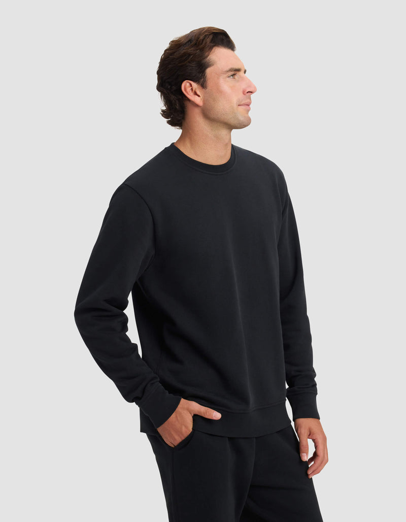A man stands against a plain light gray background wearing the Cozy Earth Men's Dylan Crewneck and matching black pants, with one hand in his pocket and a neutral expression as he looks to his left.