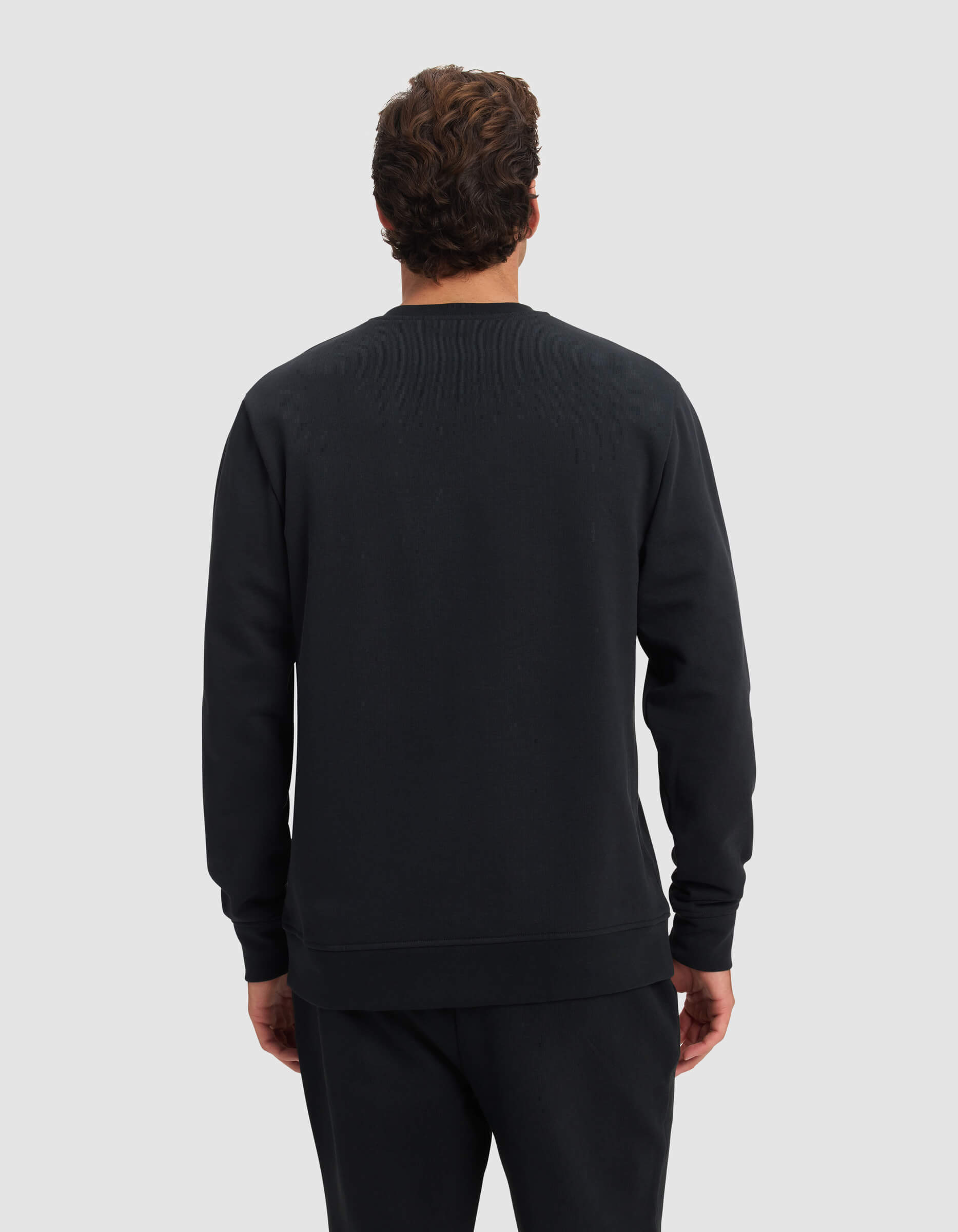 A man with curly brown hair stands facing away from the camera, wearing the Cozy Earth Men's Dylan Crewneck in black and black pants against a plain light gray background.