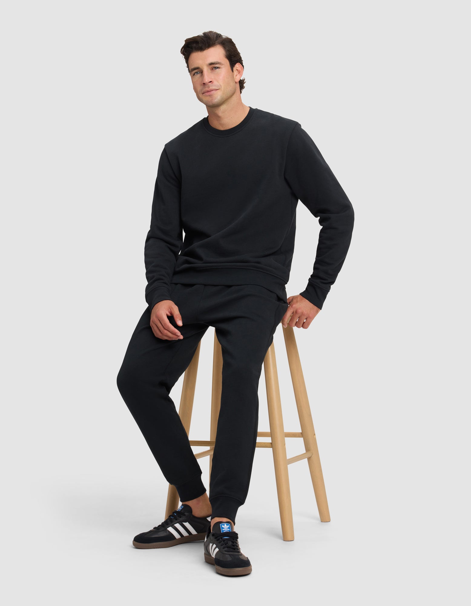 A man wears the Cozy Earth Men's Dylan Crewneck with matching black sweatpants and Adidas sneakers with white stripes, sitting on a wooden stool against a plain light gray background, looking relaxed.