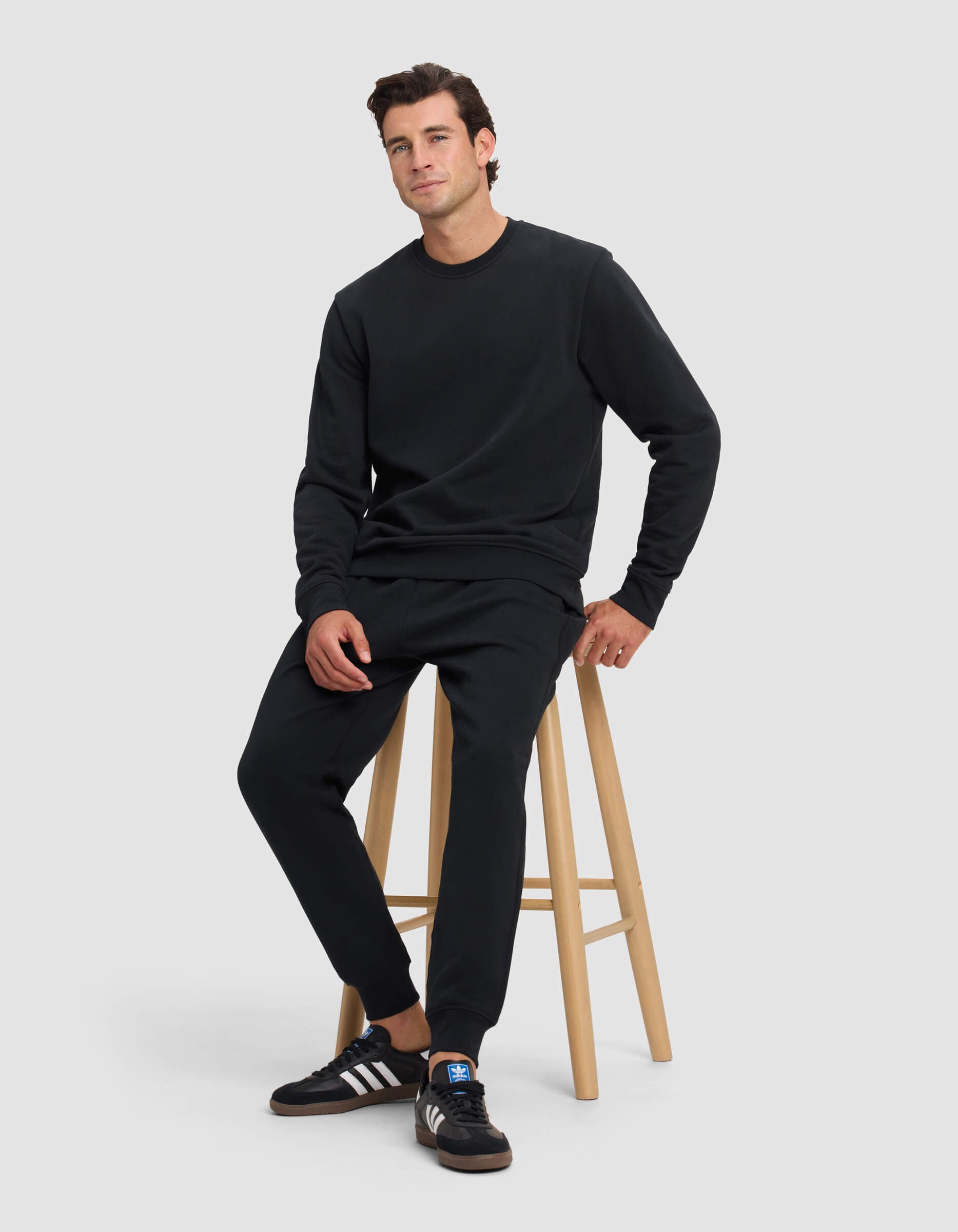 A man wearing the Cozy Earth Men's Dylan Crewneck and black pants sits on a wooden stool against a plain light gray background. He looks relaxed, with one hand on his knee, pairing his outfit with black Adidas sneakers featuring white stripes.