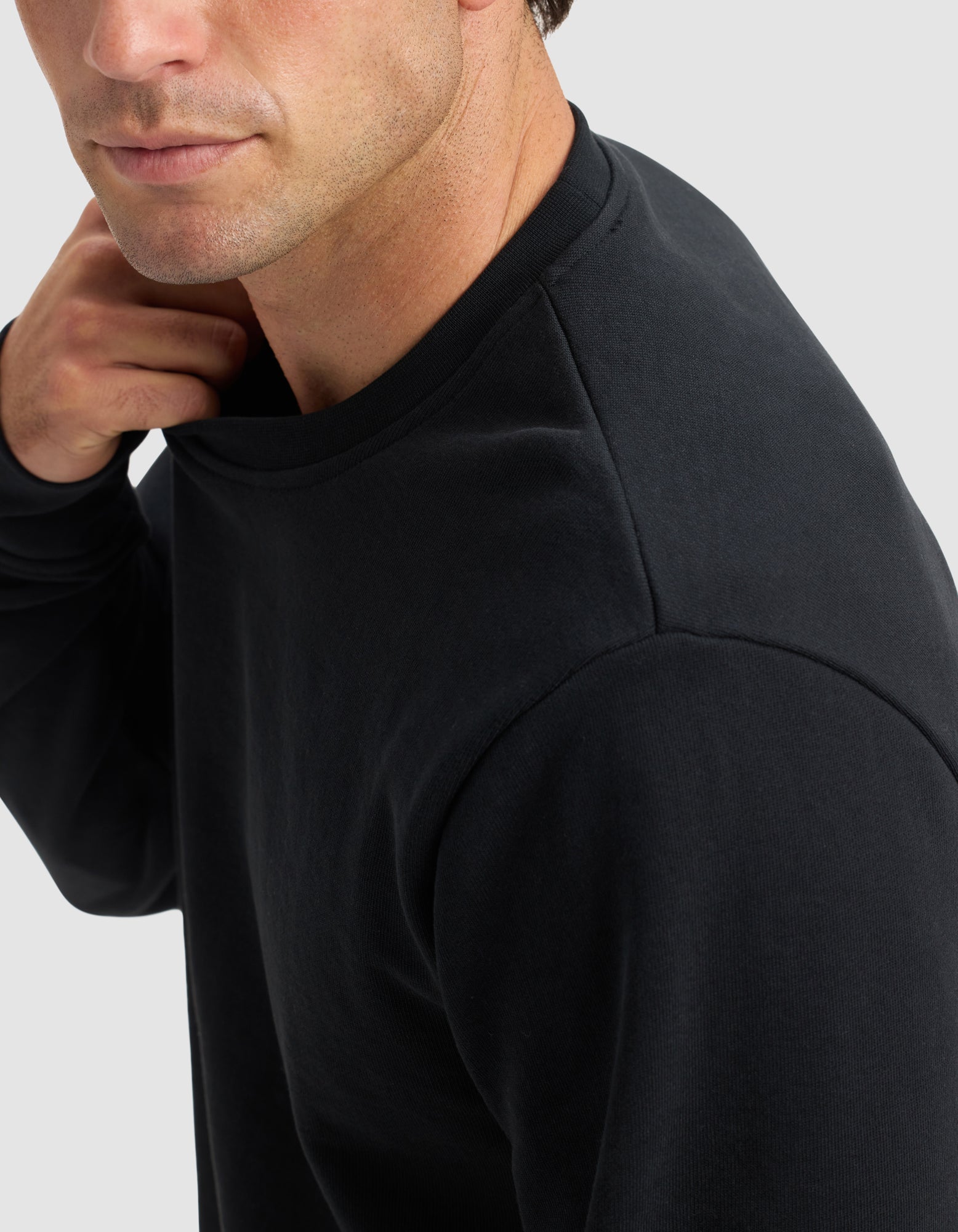 A man is shown from the shoulders up, slightly turned to the side, wearing the Men's Dylan Crewneck by Cozy Earth in black. He touches his neck against a plain light gray background.