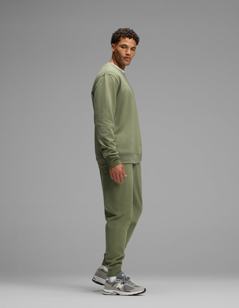 A man wearing an olive green Men’s Dylan Crewneck & Jogger Set featuring a matching crewneck sweatshirt and jogger pants.