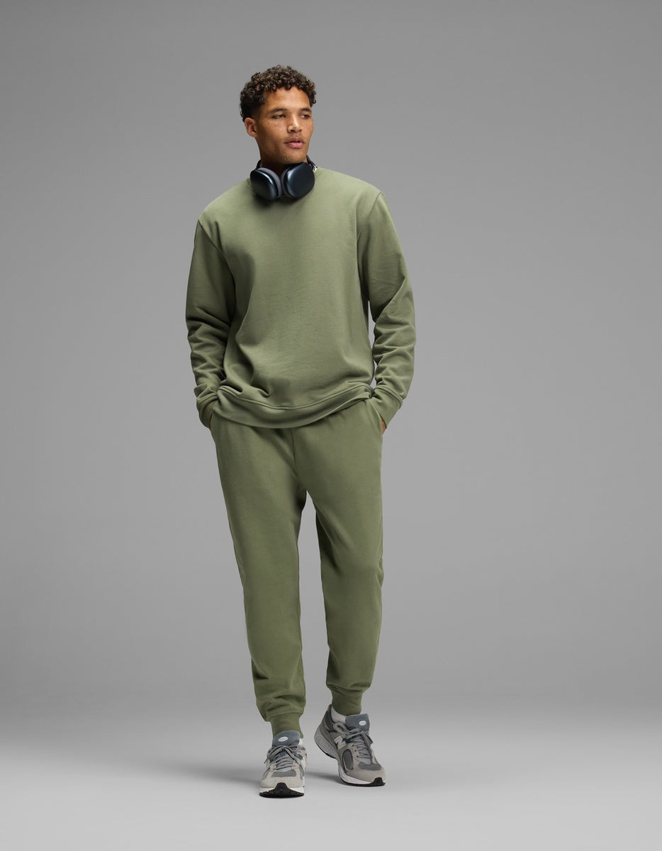 A man wearing an olive green Men’s Dylan Crewneck & Jogger Set with over-ear headphones around his neck.