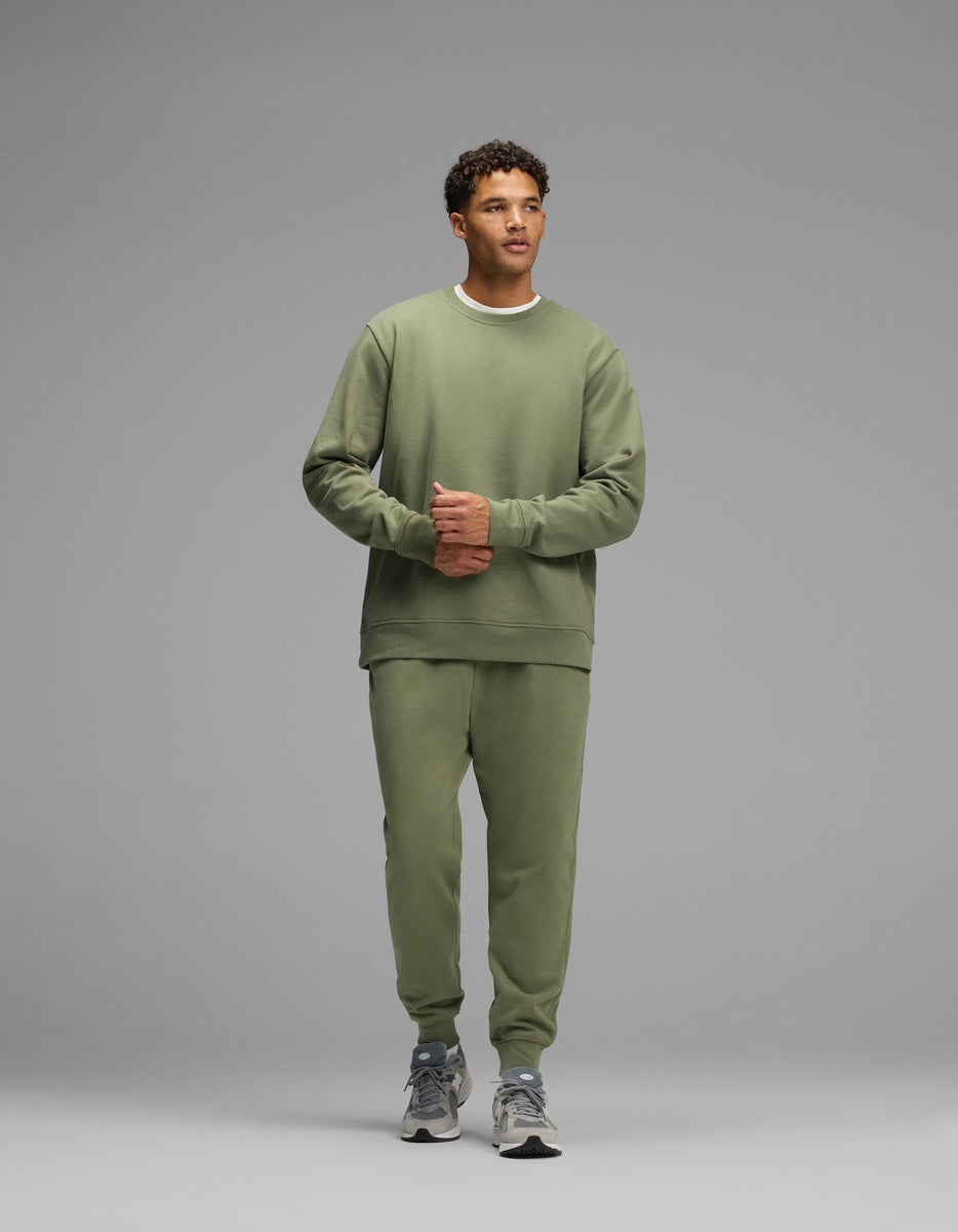 A man wears an olive green Cozy Earth Men’s Dylan Crewneck and matching sweatpants with grey sneakers.
