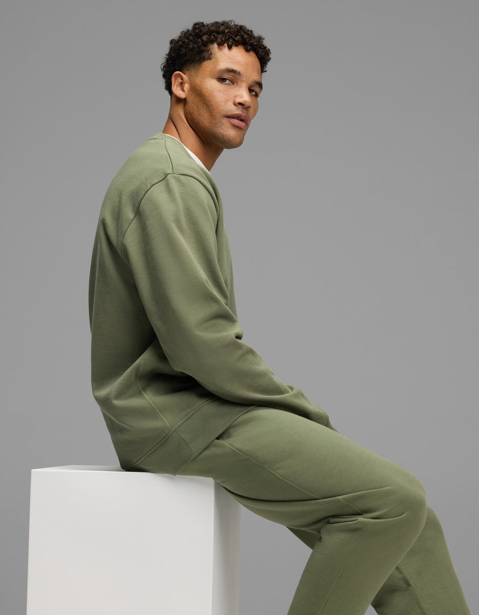 A man wearing a matching olive green Cozy Earth Men’s Dylan Crewneck and sweatpants, sitting on a white pedestal.