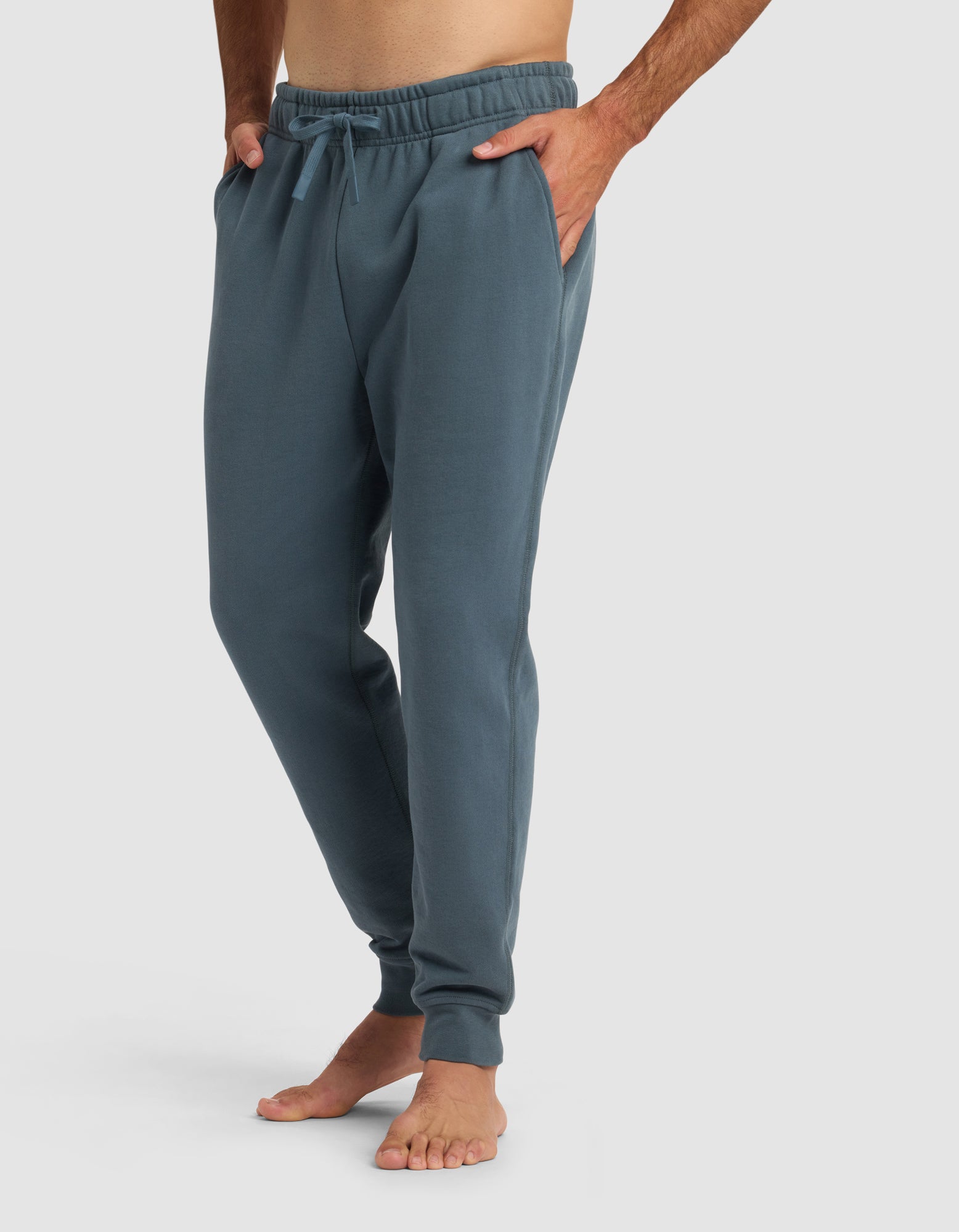 A barefoot man wears the Cozy Earth Men's Dylan Jogger, featuring an elastic waistband and drawstring, as he stands with hands in his pockets against a plain light background.