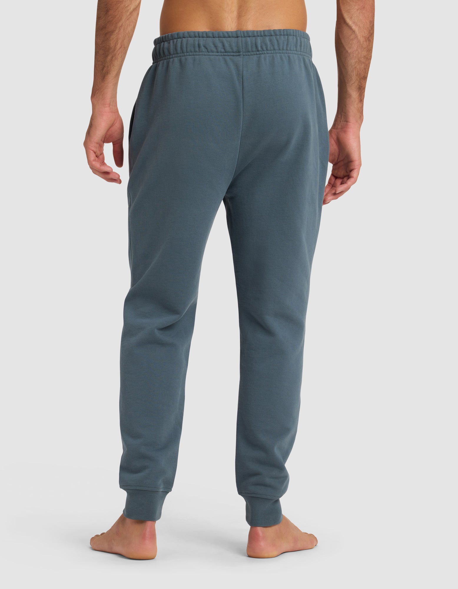 A barefoot man is seen from behind wearing Cozy Earth's Men's Dylan Jogger in blue-gray, featuring an elastic waistband and cuffed ankles, standing against a plain white background.