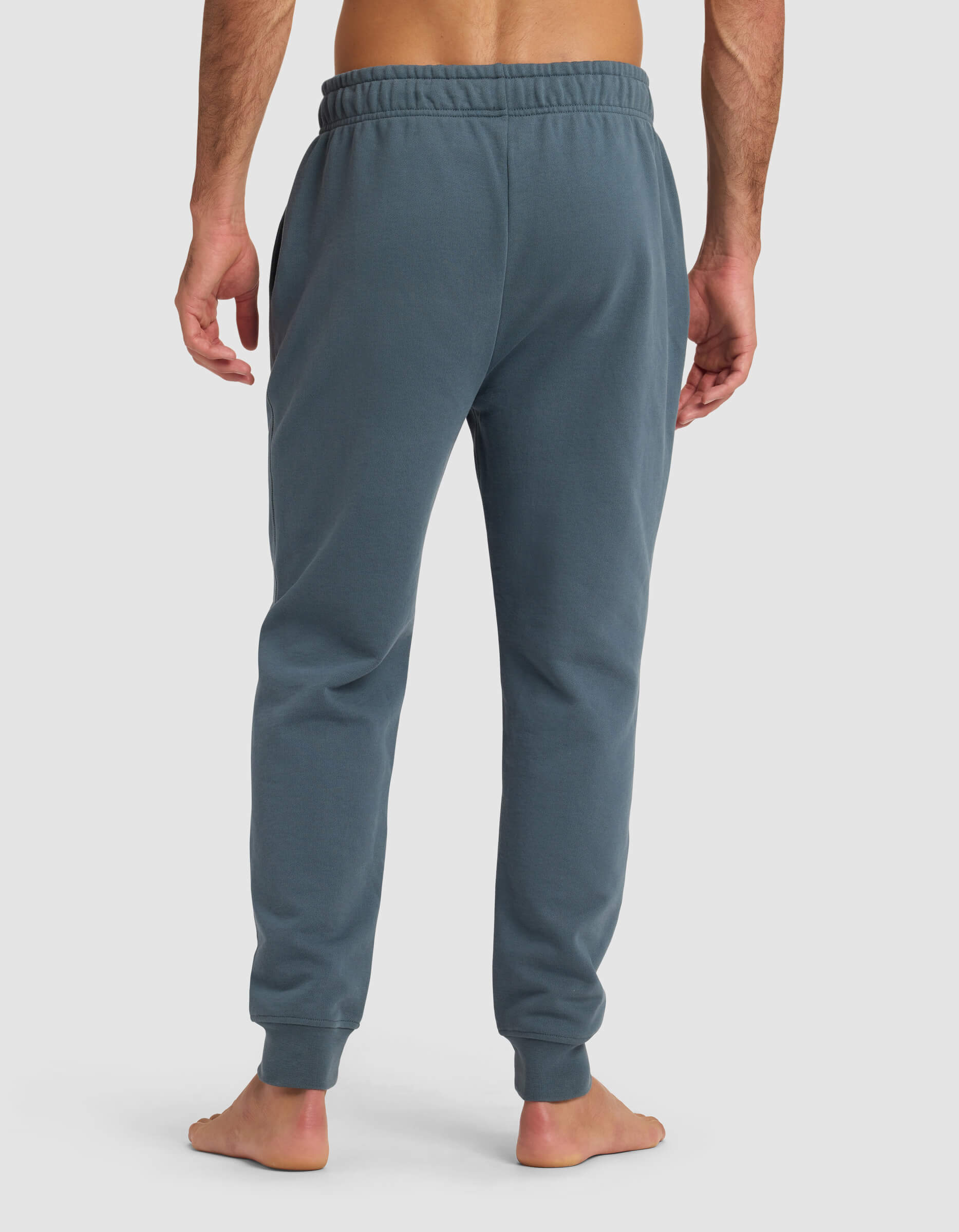 A barefoot man faces away, wearing Cozy Earth Men's Dylan Jogger in blue-green with an elastic waistband and cuffed ankles, set against a plain light background.