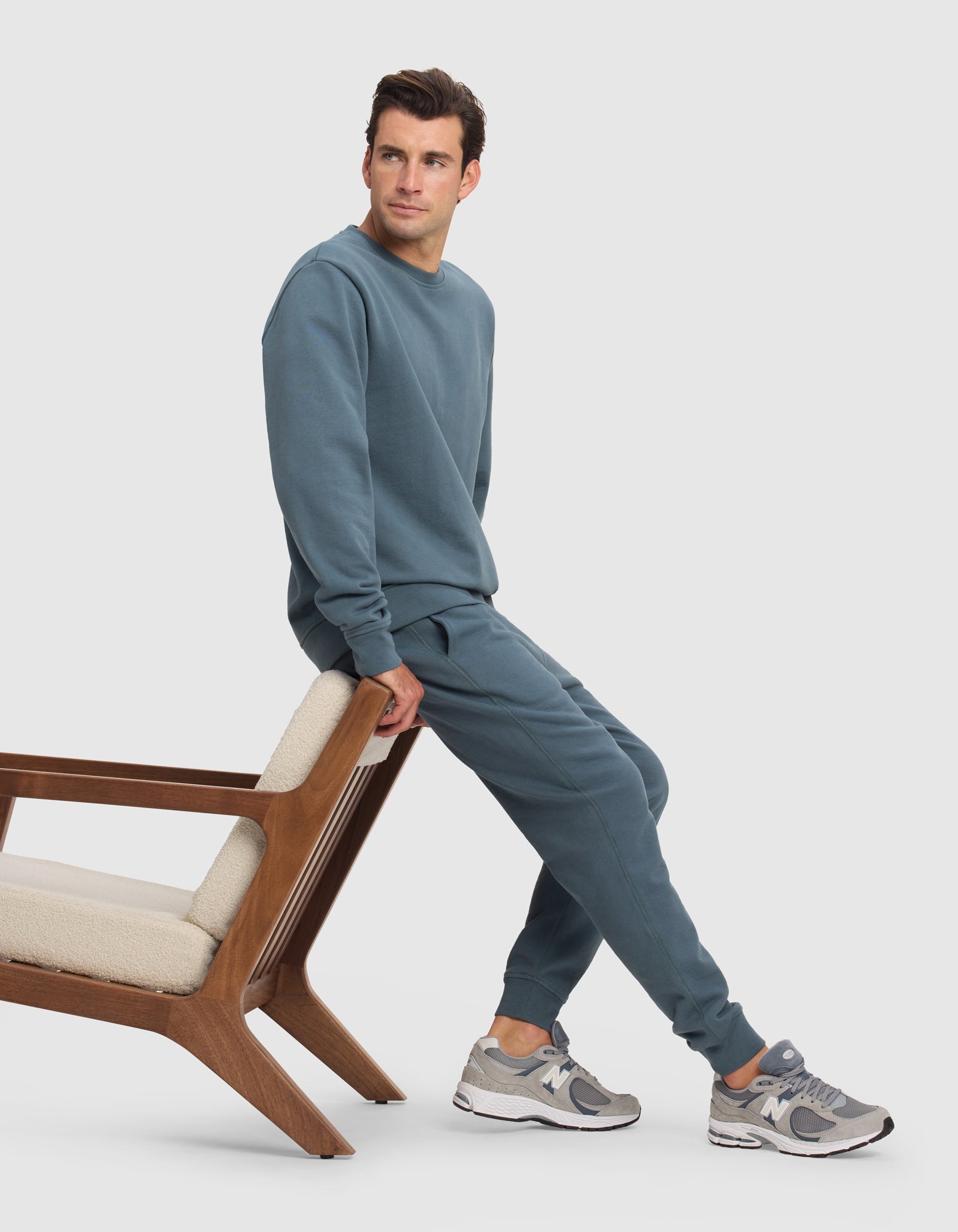 A man sits on a modern wooden chair, looking to the side, wearing Cozy Earth’s Men's Dylan Jogger in blue with a matching sweatshirt and gray New Balance sneakers. The background is plain white.|Color:Dawn