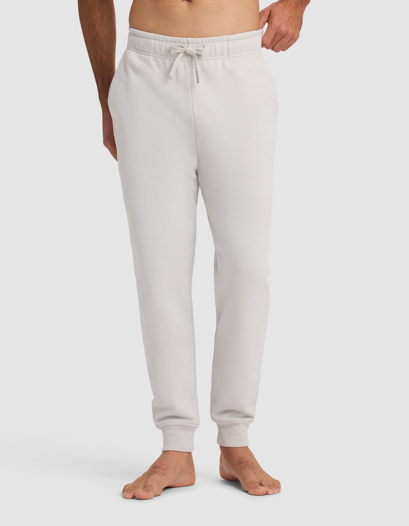 A man stands barefoot against a white background wearing Cozy Earth's Men's Dylan Jogger in light gray, featuring an elastic waistband, drawstring, and side pockets.