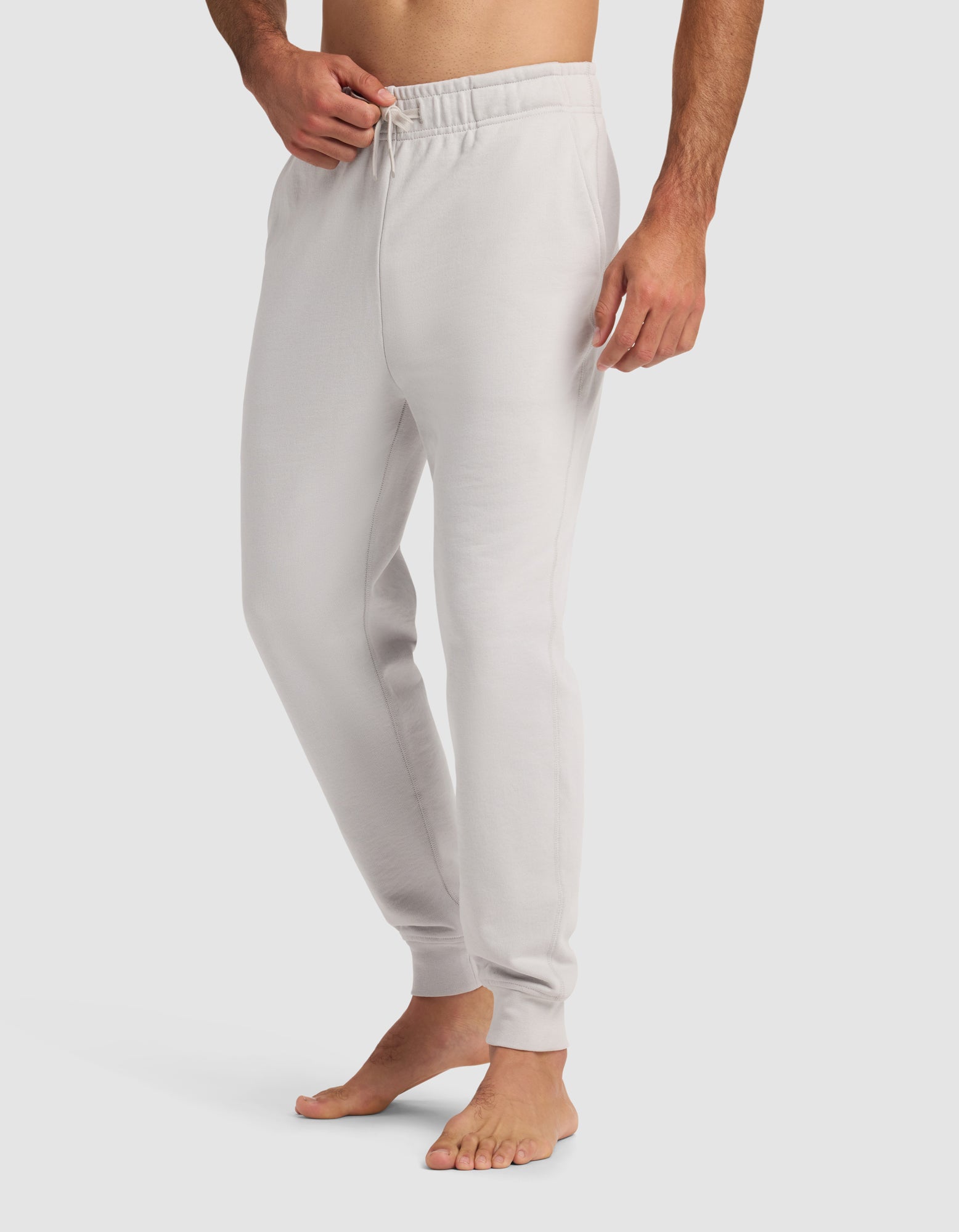 A barefoot man models the Cozy Earth Men's Dylan Jogger—light gray, fitted sweatpants with an elastic waistband and drawstring—against a plain white background. Only his lower body is visible.