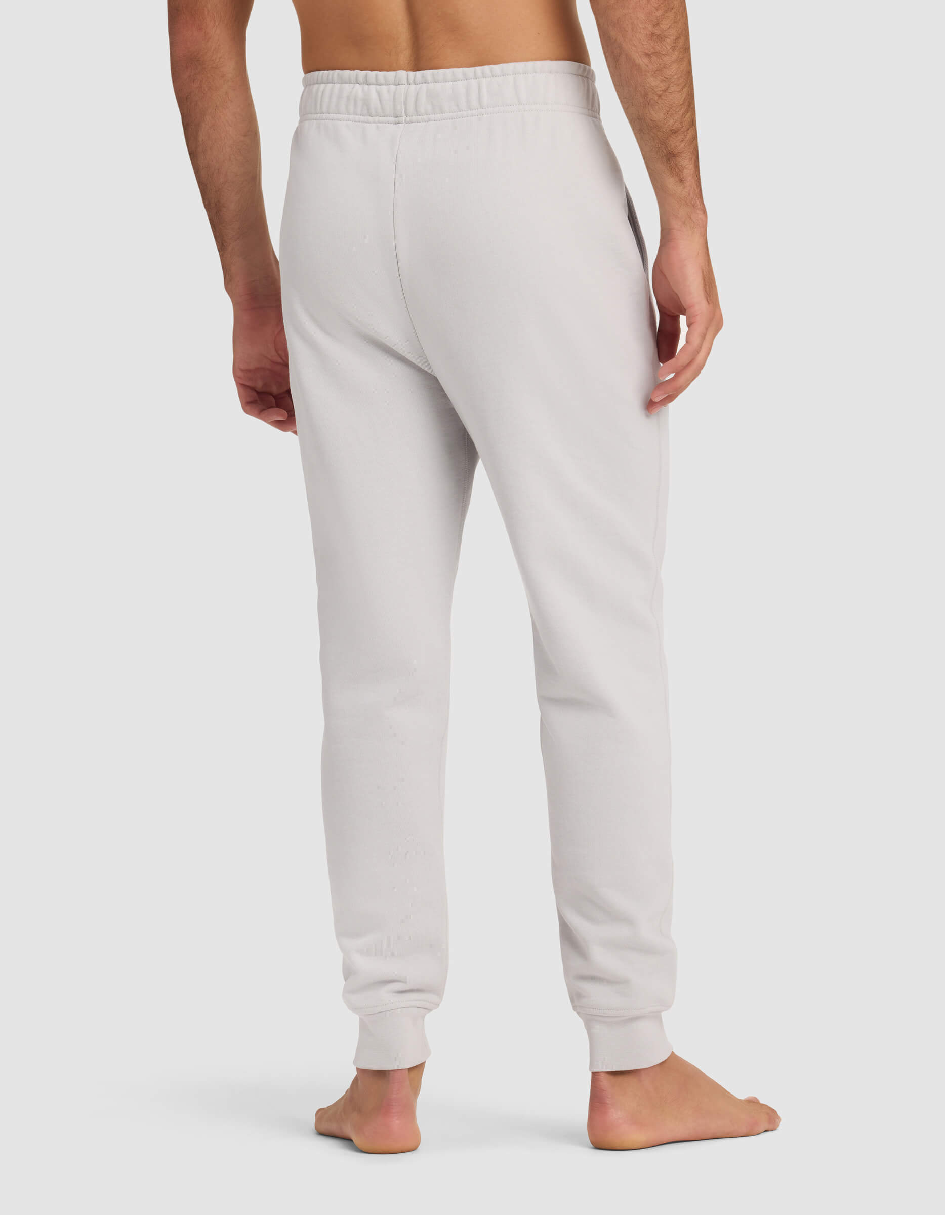 A barefoot man is shown from the back wearing Cozy Earth's Men's Dylan Jogger in light gray, featuring an elastic waistband and cuffed ankles, standing against a plain white background.|Color:Dew