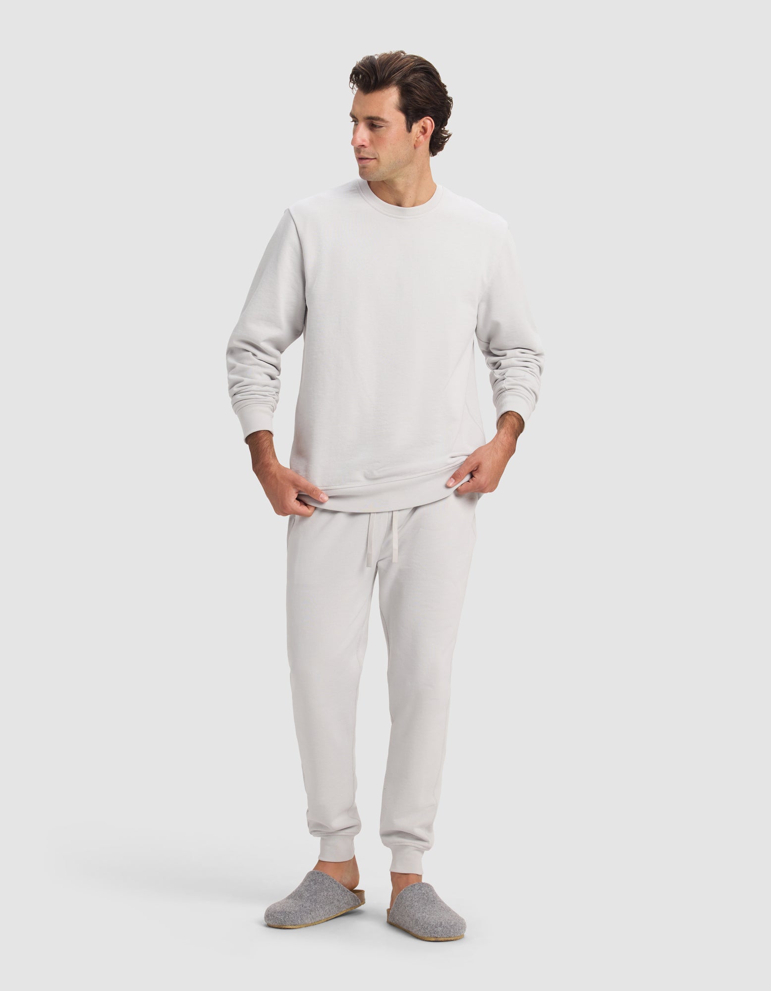 A man wears the Cozy Earth Men's Dylan Jogger in light gray with a matching sweatshirt and slippers, adjusting his sweatshirt hem as he looks left, posing against a plain light background.