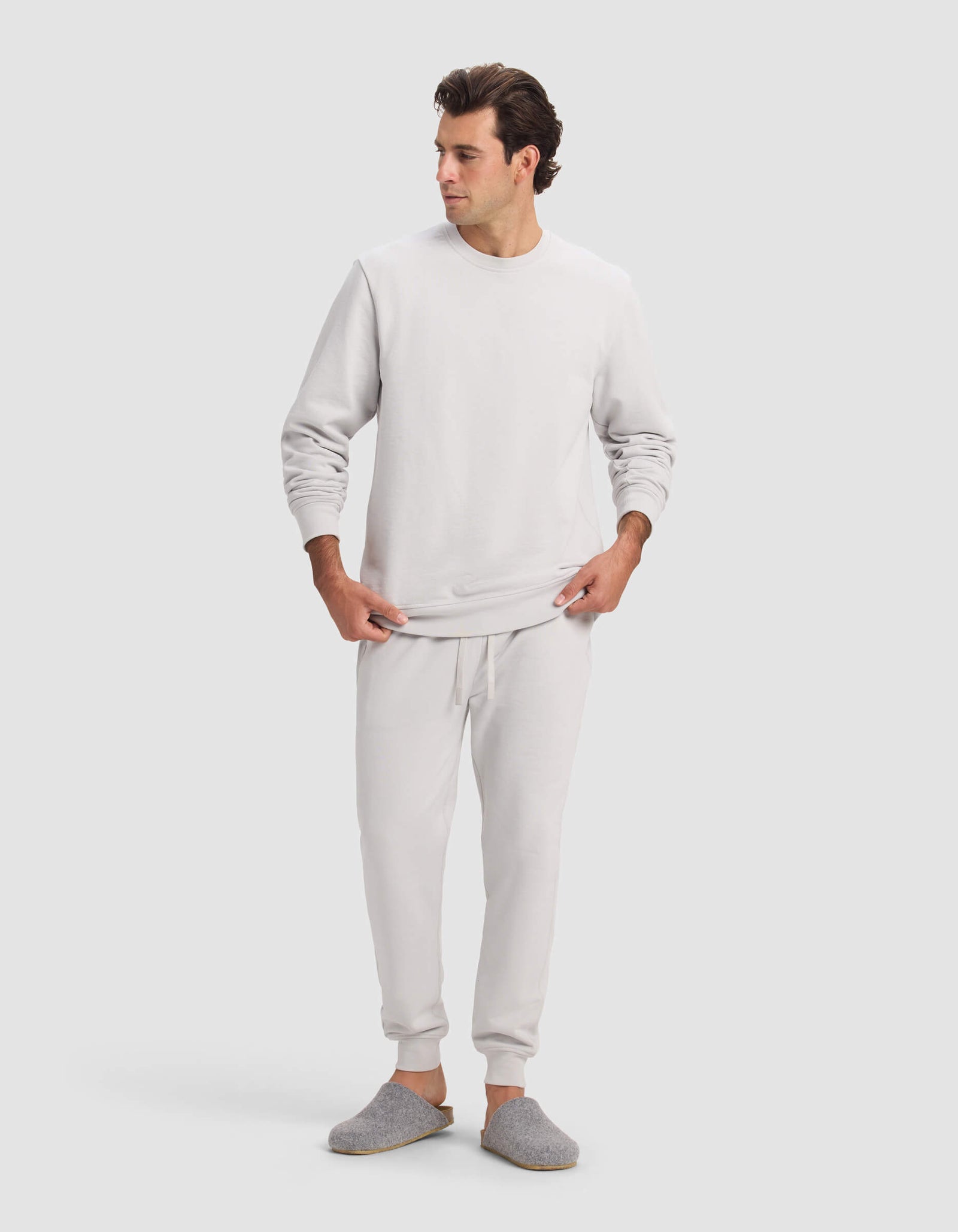 A man wears the Cozy Earth Men's Dylan Jogger in light gray with matching joggers and slippers, standing against a plain, light background, looking to his left with hands near his waist.
