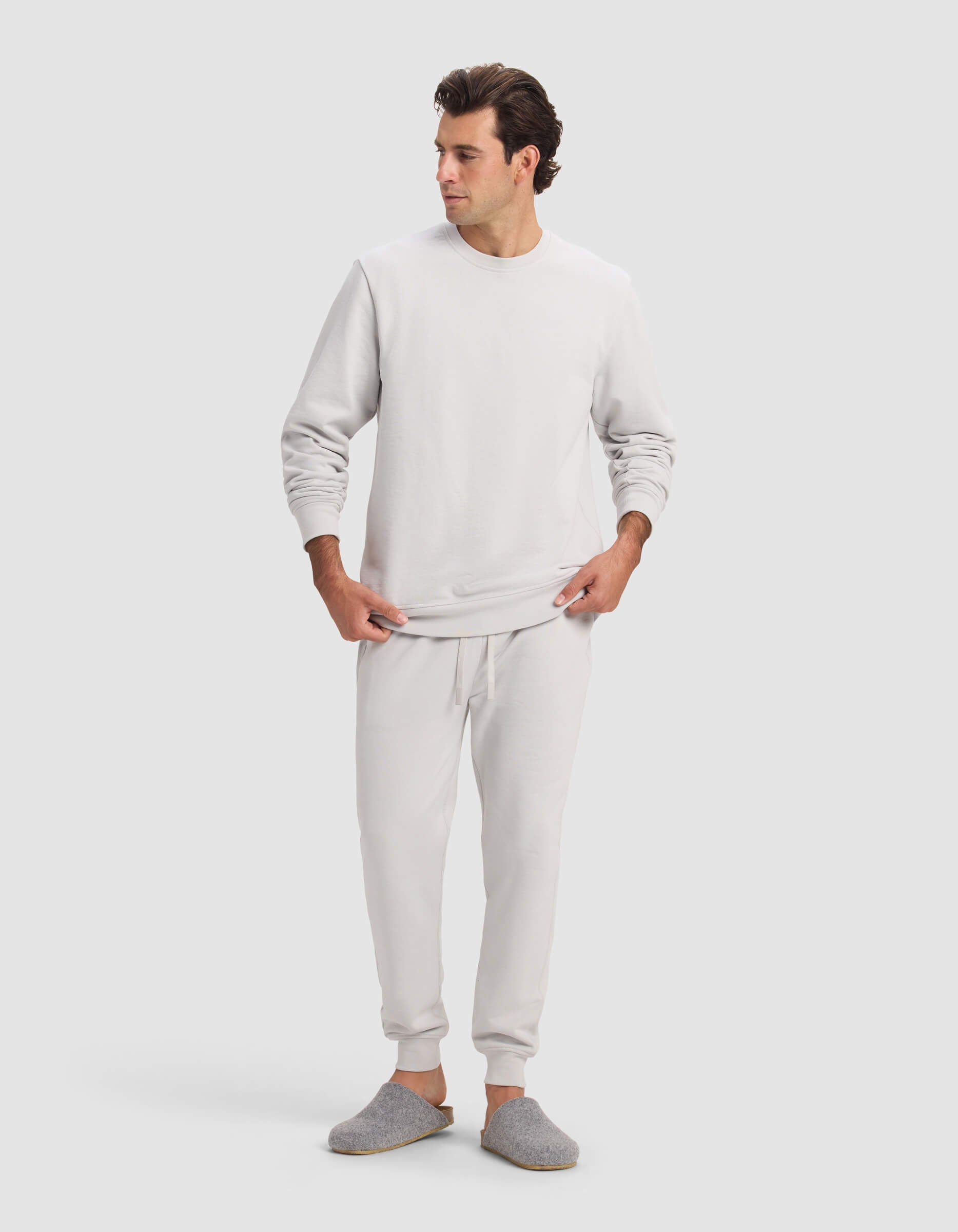 A man wears the Cozy Earth Men's Dylan Jogger in light gray with matching joggers and slippers, standing against a plain, light background, looking to his left with hands near his waist.|Color:Dew