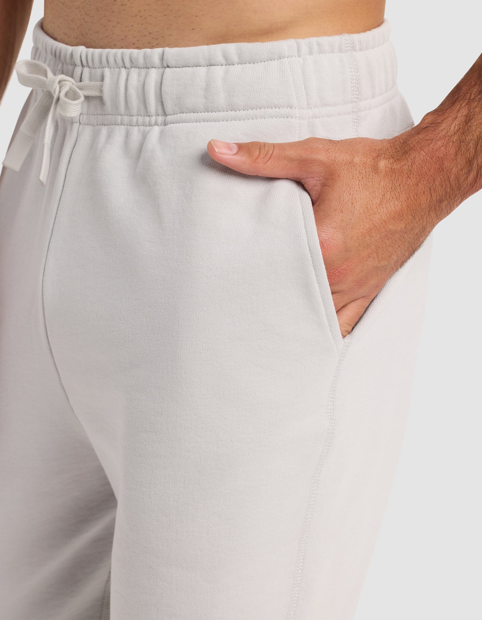 A man wears Cozy Earth's Men's Dylan Jogger in light gray, featuring a drawstring waistband and pockets, standing with one hand in his pocket for a close-up view of the pants and hand.