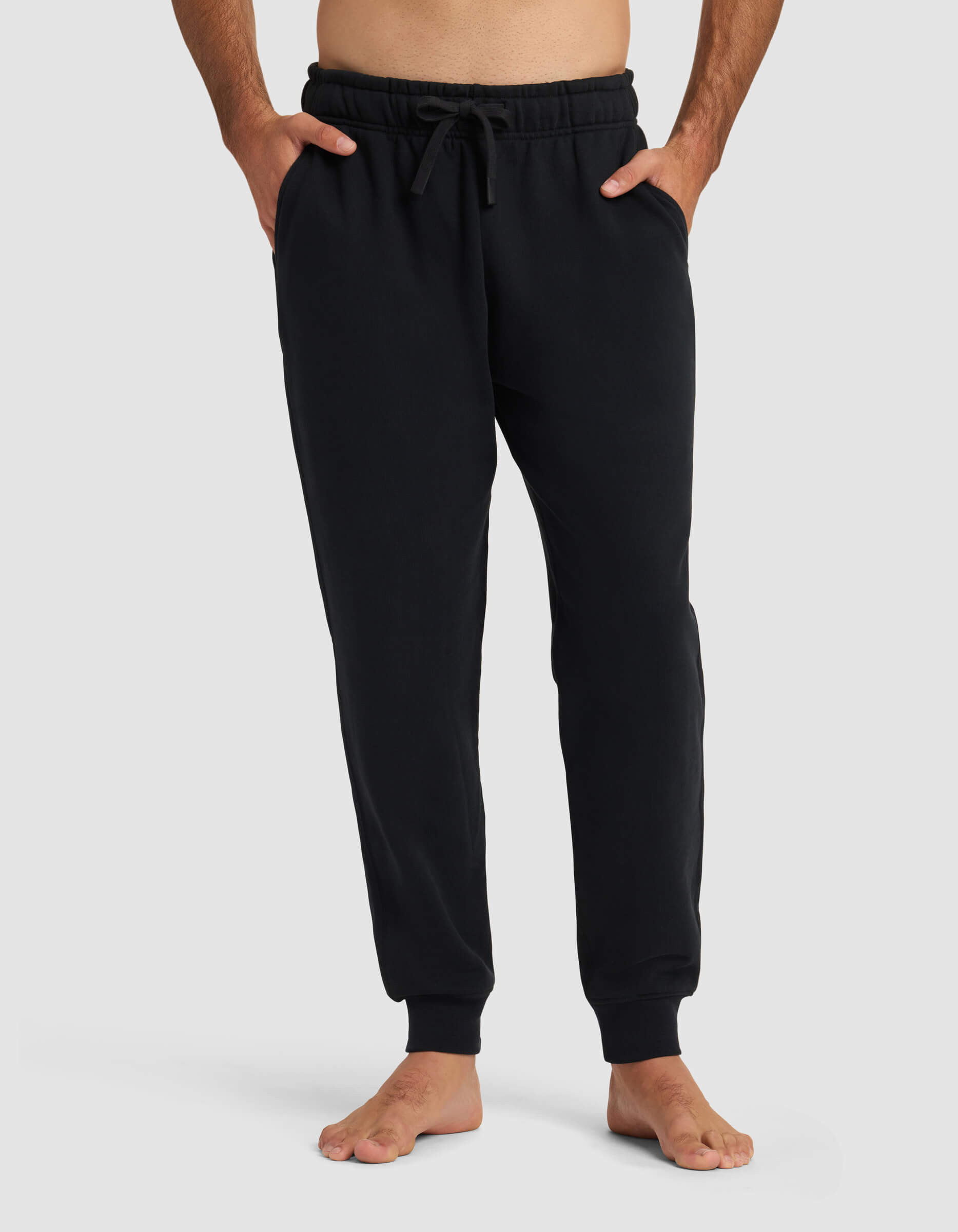 A person stands barefoot in Cozy Earth Men's Dylan Jogger in Jet Black, featuring an elastic waistband, drawstring, and side pockets. The background is plain light gray.|Color:Jet Black