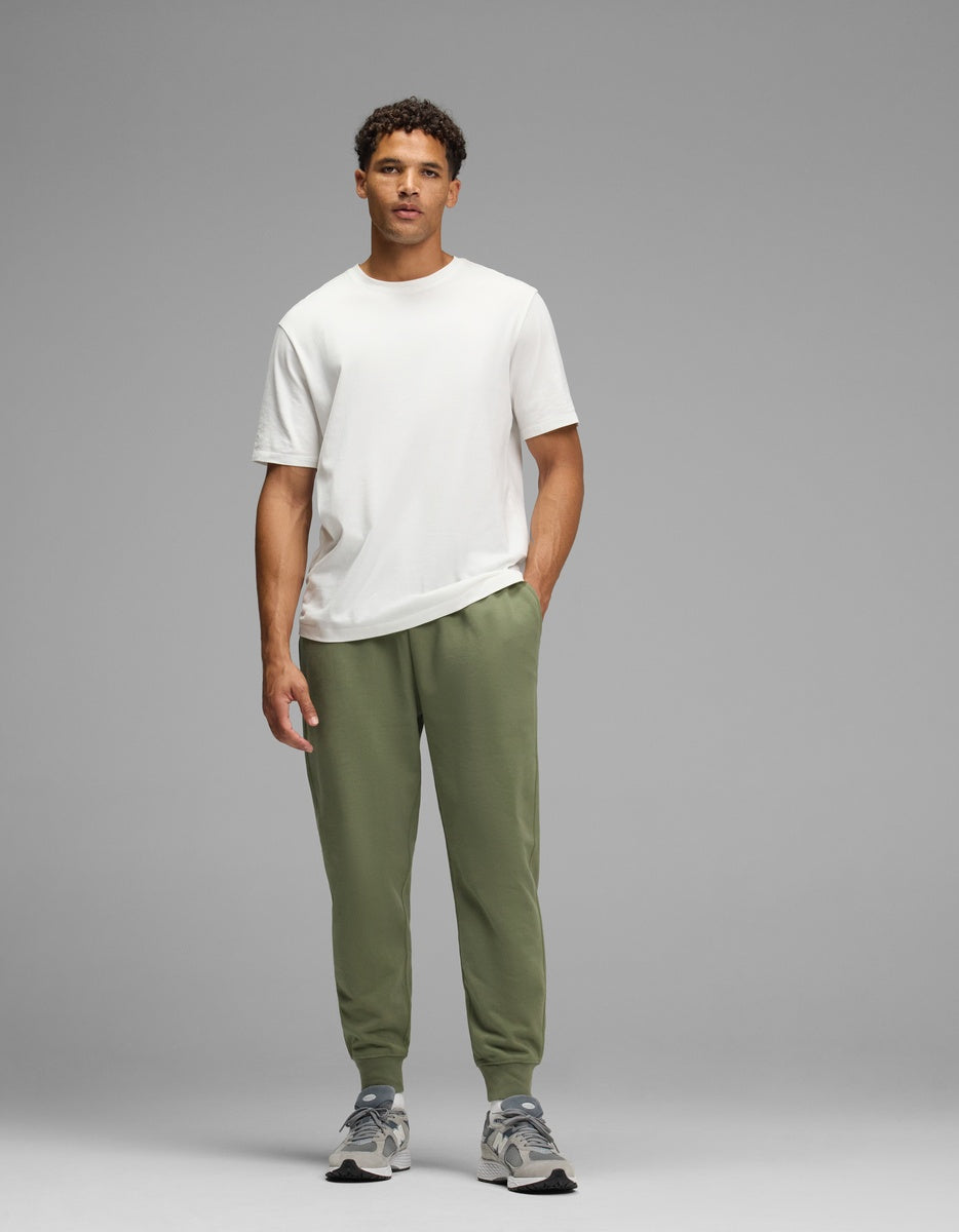 A man wears a white t-shirt, olive green Men’s Dylan Jogger pants with cuffed ankles, and grey athletic shoes.