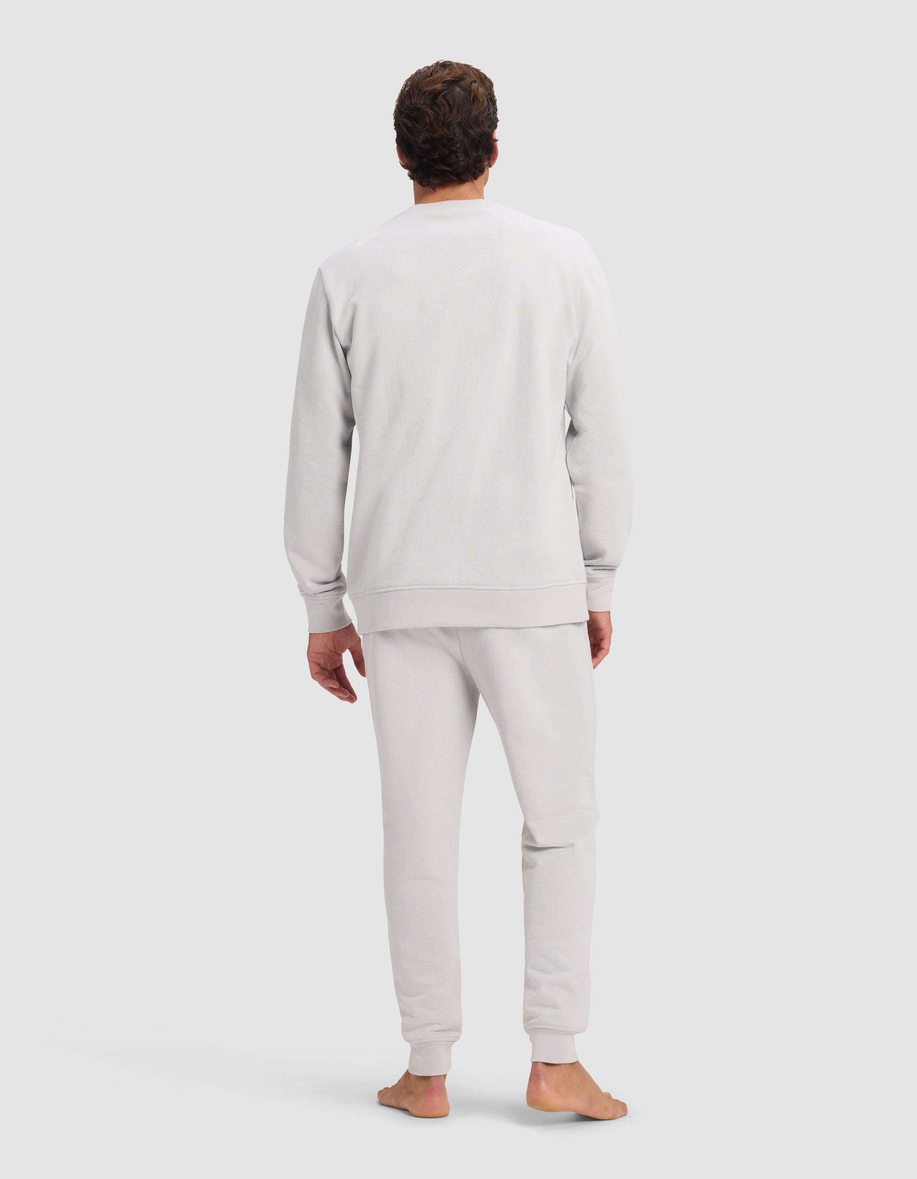 A person stands barefoot facing away, wearing the Cozy Earth Men's Dylan Crewneck & Jogger Set in light gray against a plain light gray background.