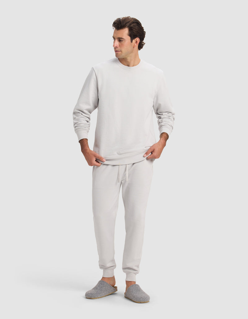A man wears the Cozy Earth Men's Dylan Jogger in light gray with matching joggers and slippers, standing against a plain, light background, looking to his left with hands near his waist.