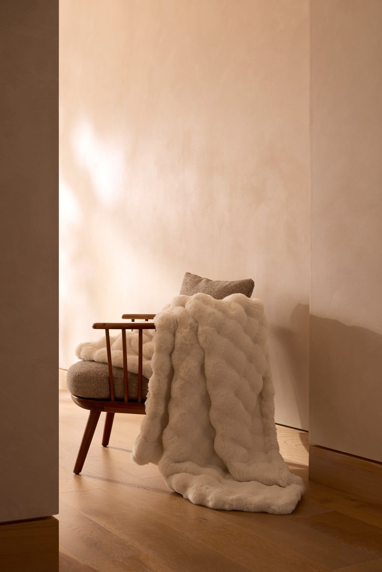 A wooden chair with a light cushion and a Cozy Earth Bubble Cuddle Blanket in plush white faux fur sits on a wooden floor against a beige wall in a softly lit, minimalist room. |Color:Creme