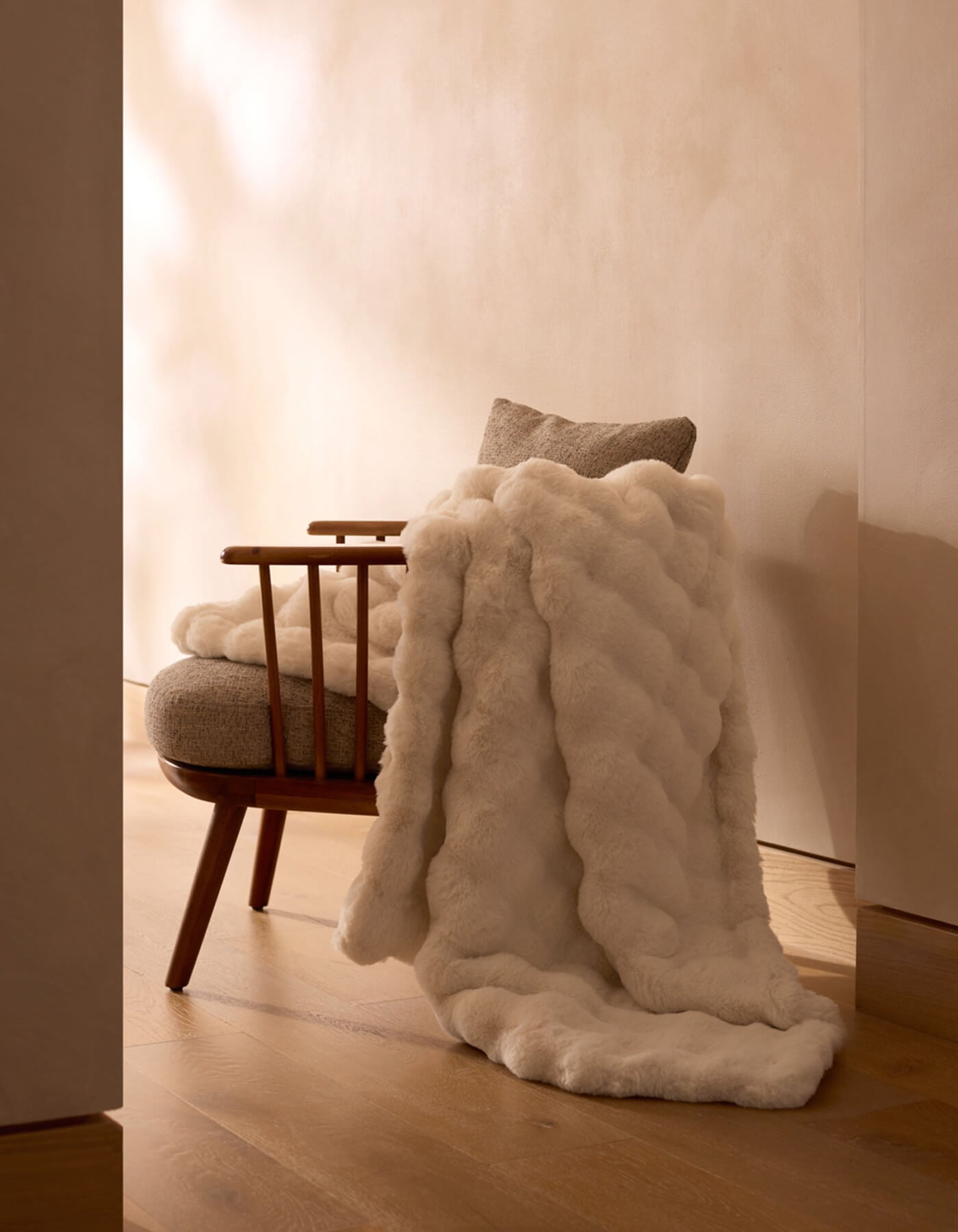 A wooden chair with a light cushion and a Cozy Earth Bubble Cuddle Blanket in plush white faux fur sits on a wooden floor against a beige wall in a softly lit, minimalist room. |Color:Creme