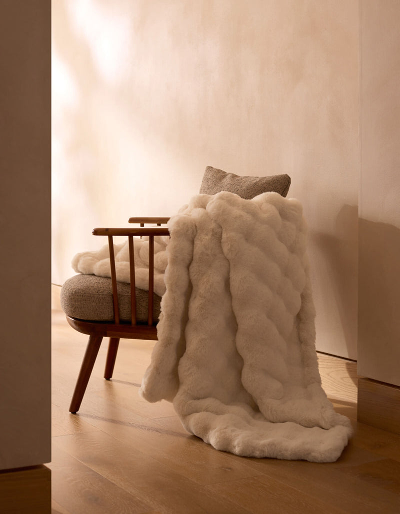 A wooden chair with a light cushion and a Cozy Earth Bubble Cuddle Blanket in plush white faux fur sits on a wooden floor against a beige wall in a softly lit, minimalist room.