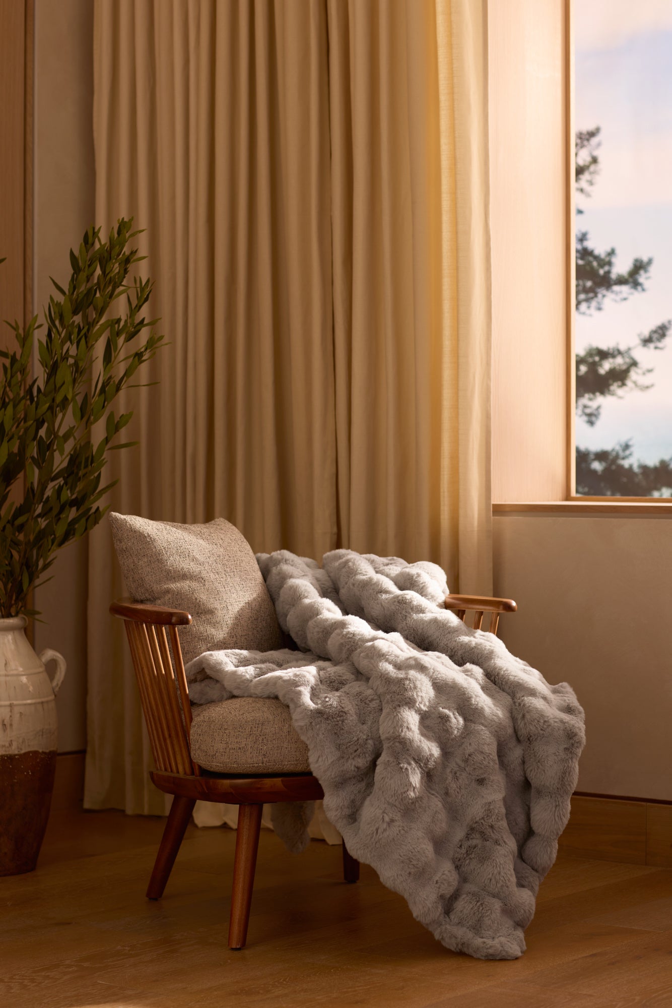A beige armchair features the Bubble Cuddle Blanket by Cozy Earth draped over it. Sunlight pours through cream curtains, lighting up the wooden floor and a potted plant in a ceramic vase nearby. 