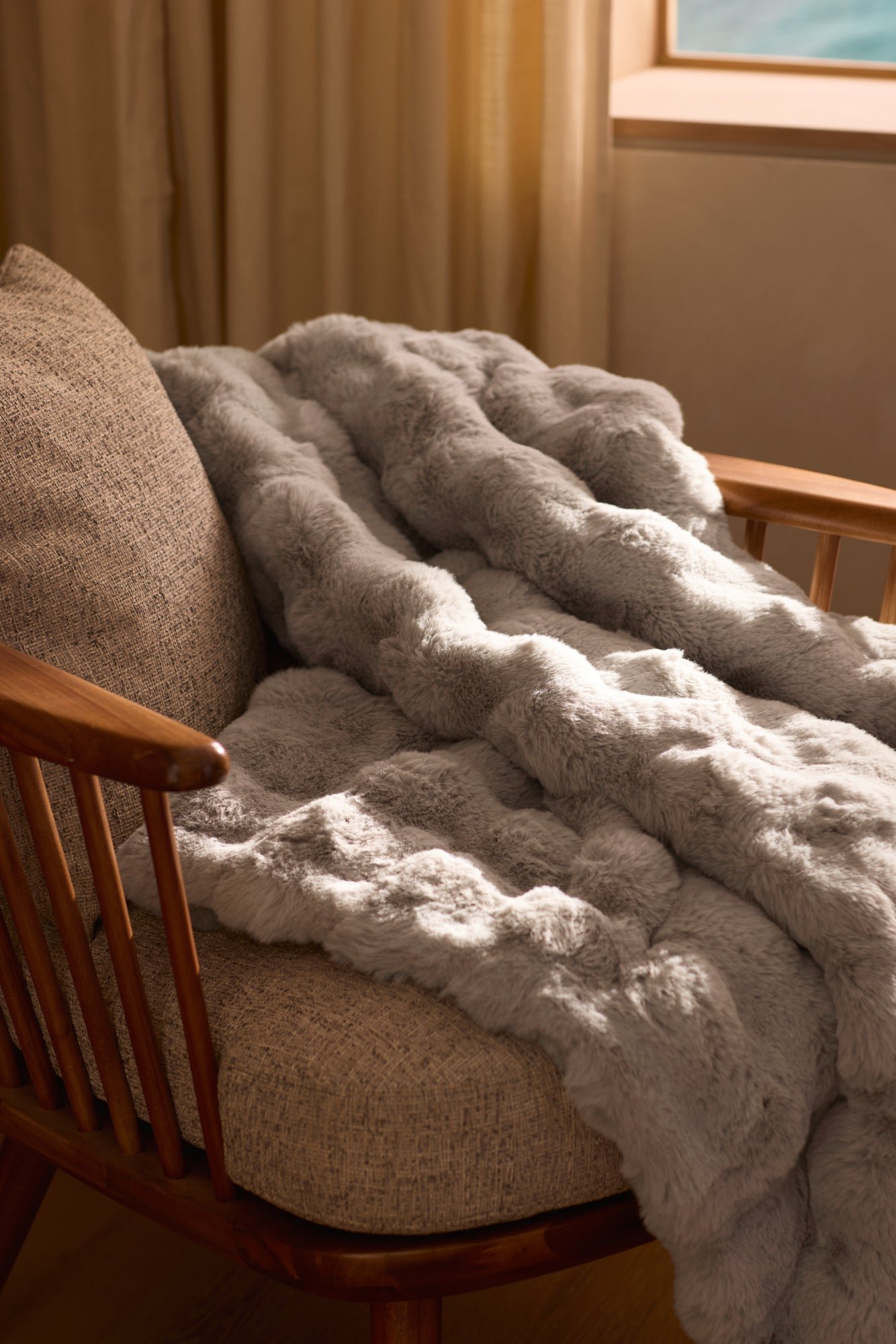 The Cozy Earth Bubble Cuddle Blanket, in plush light gray faux fur, is draped over a cushioned wooden chair by a window with beige curtains, adding warmth and inviting comfort to the room. |Color:Light Grey