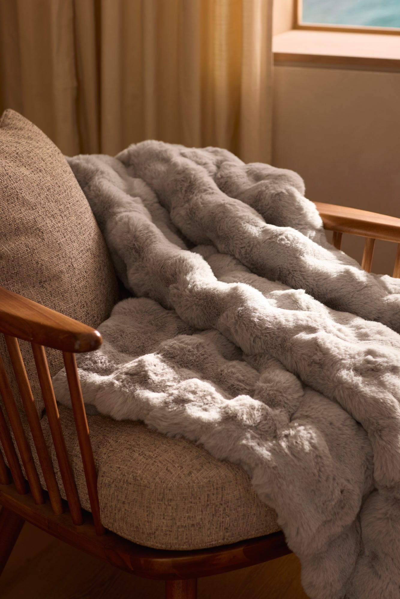 The Cozy Earth Bubble Cuddle Blanket, in plush light gray faux fur, is draped over a cushioned wooden chair by a window with beige curtains, adding warmth and inviting comfort to the room. 