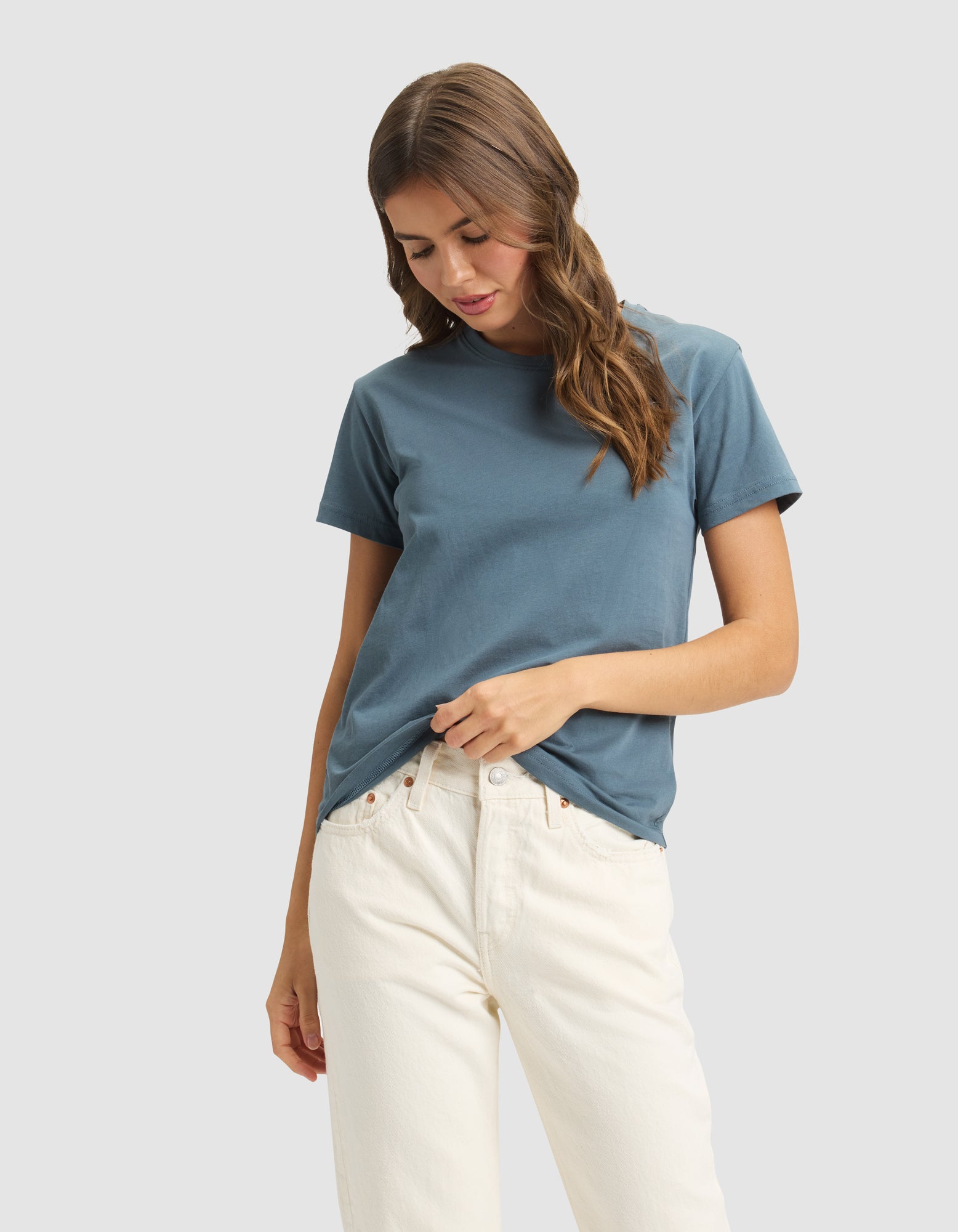 A woman with wavy brown hair wears the Cozy Earth Women's Easy Tee in blue and off-white jeans, gently holding the shirt's hem against a light gray background. 